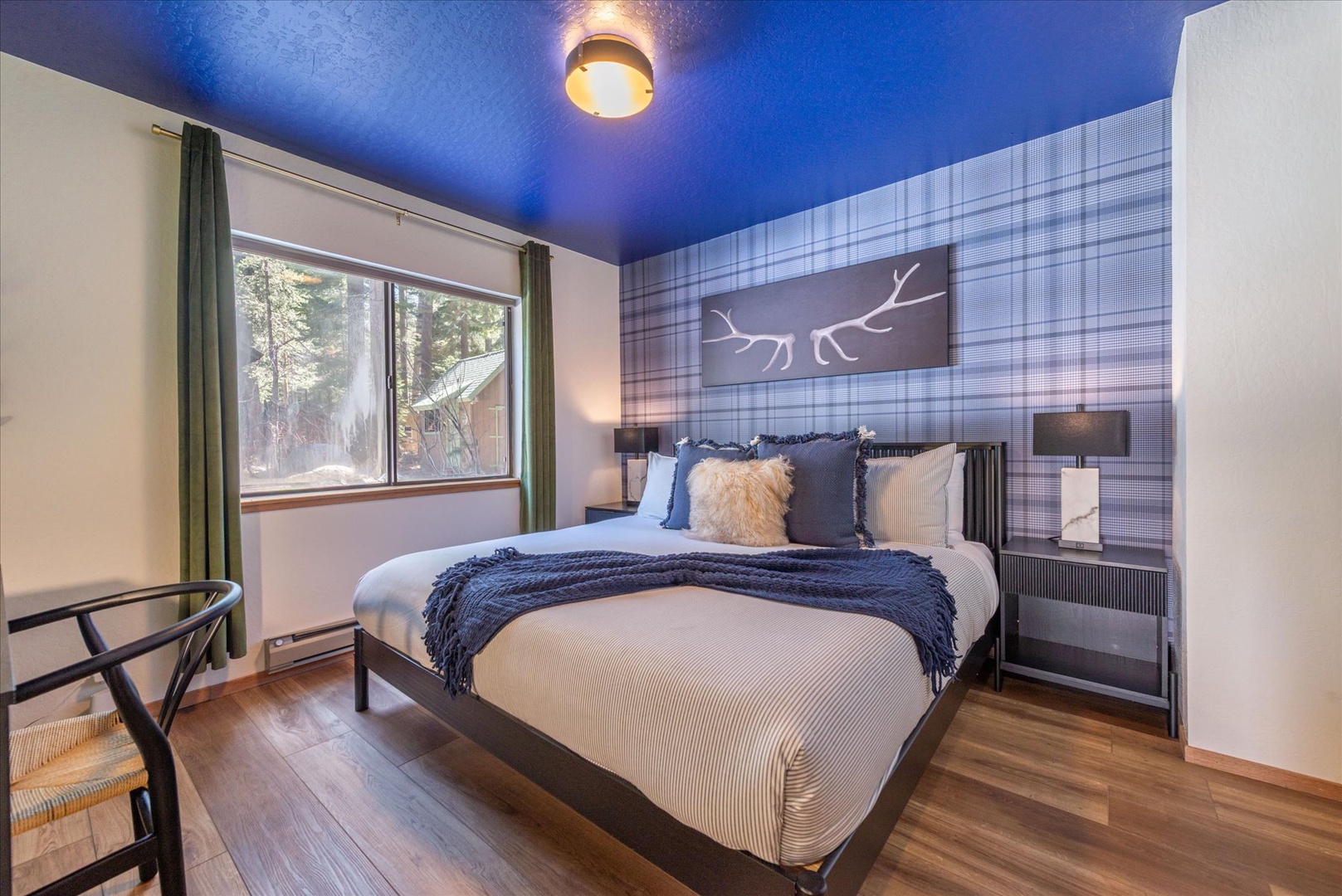 Rest peacefully in your cozy king bedroom featuring a striking blue ceiling and forest views through the window.