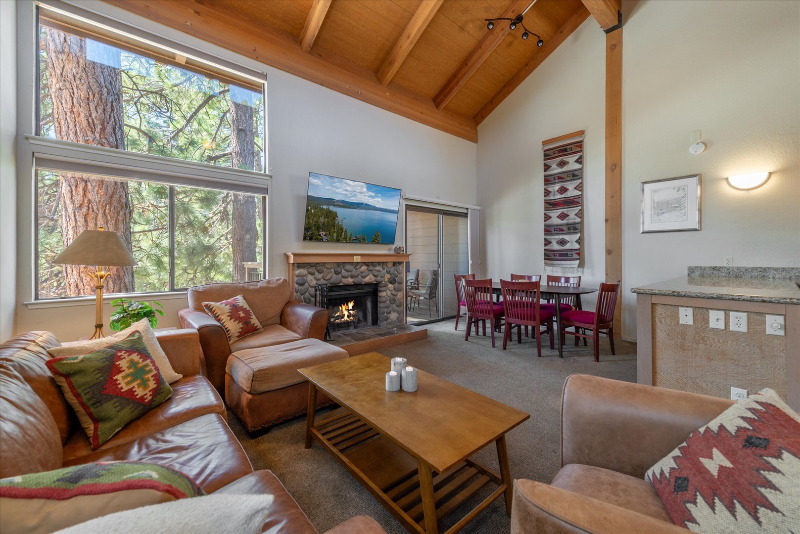 Your mountain retreat features vaulted wood-beam ceilings, a cozy stone fireplace, and large windows framing tall pines—inviting you to unwind in comfort and warmth.