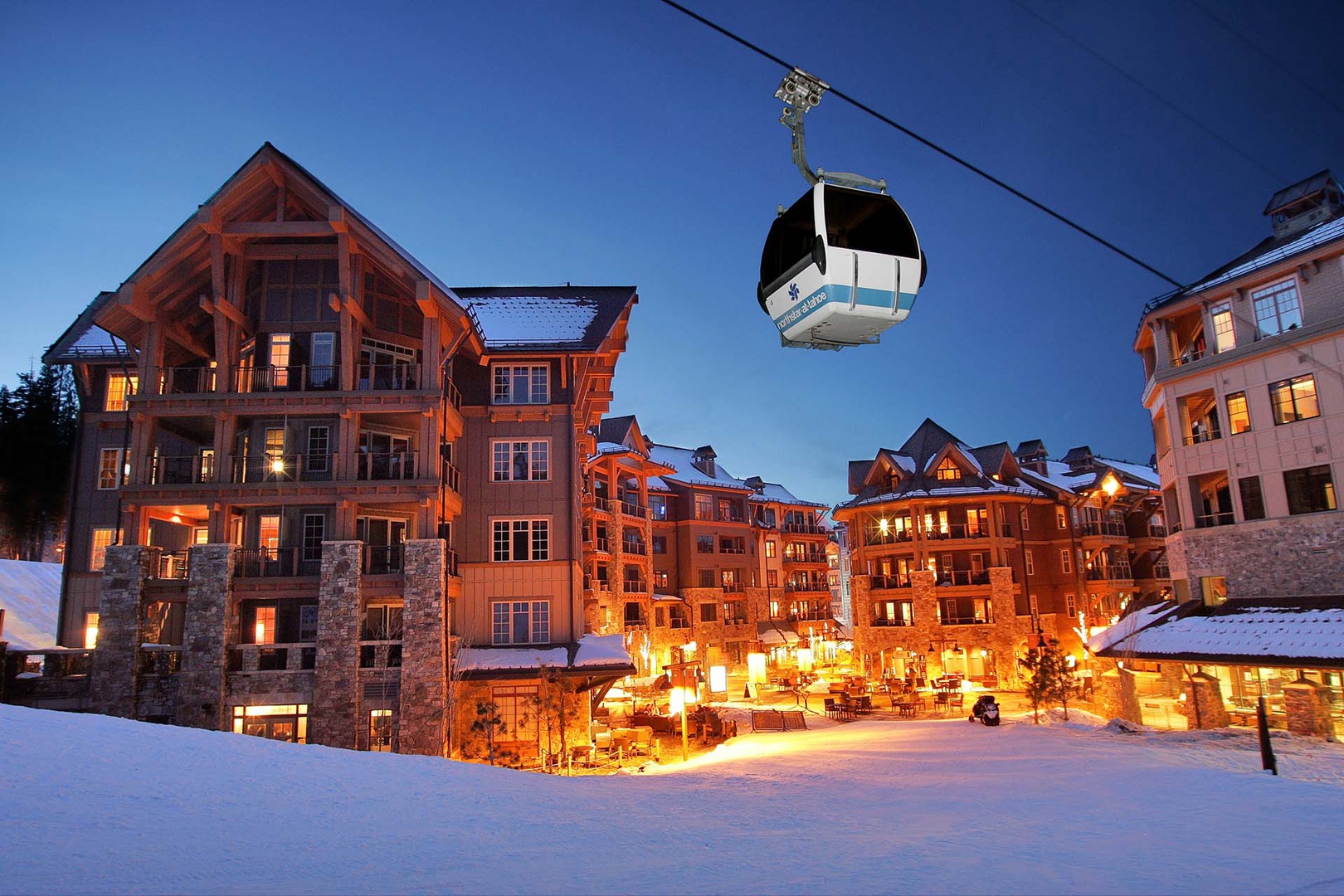 Alpine village resort with ski gondola access and illuminated mountain lodges under twilight sky.