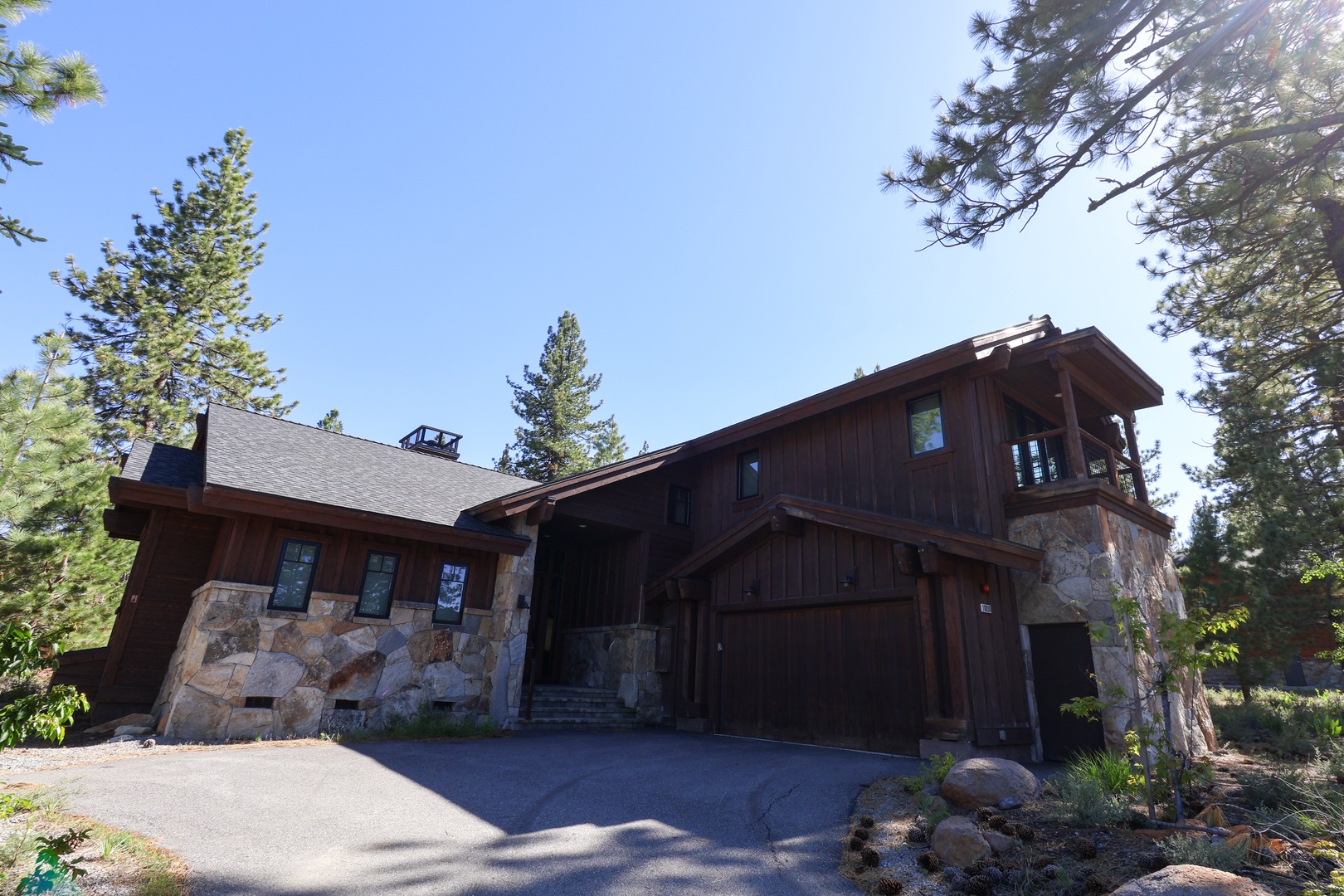 Rustic mountain retreat nestled among towering pines with stone and wood exterior, blending seamlessly into the natural forest surroundings. Spacious two-car garage provides convenient access.
