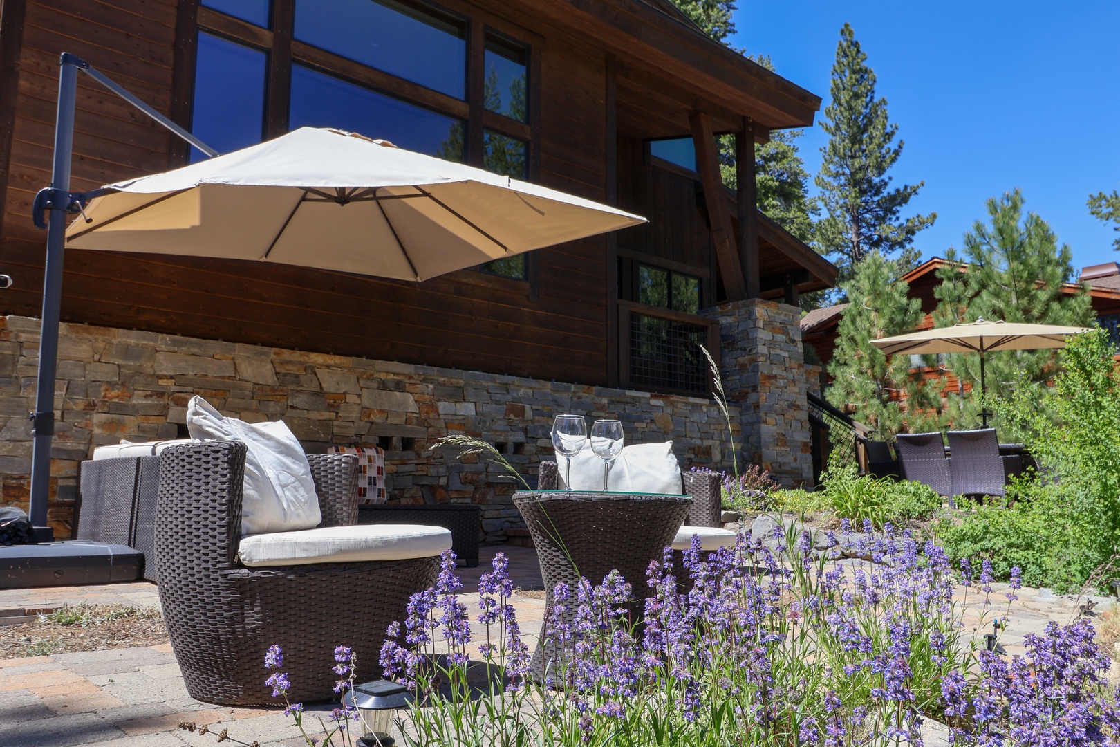 Modern mountain retreat with a stone-accented exterior set among tall pines and natural landscaping, featuring comfortable outdoor seating areas under shaded umbrellas.