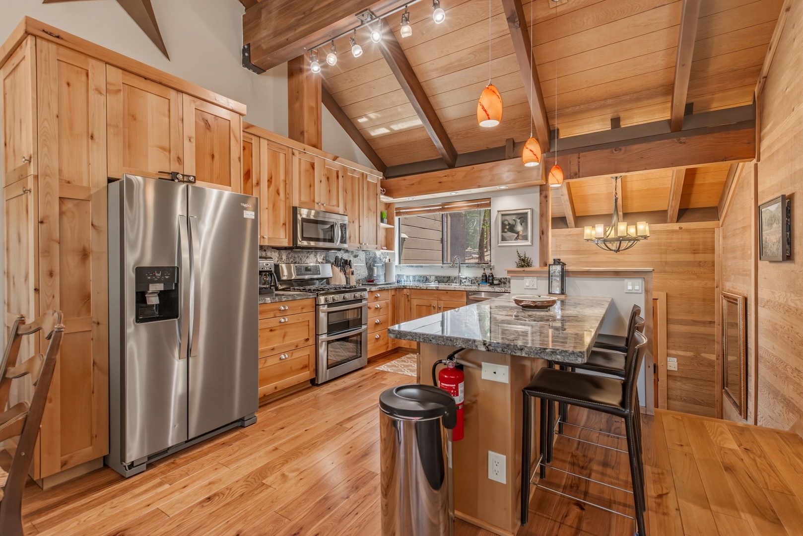 Gather around your fully equipped kitchen featuring natural wood cabinetry, granite countertops, stainless steel appliances, and an inviting breakfast bar—perfect for preparing meals together under soaring vaulted ceilings with rustic wood beams.