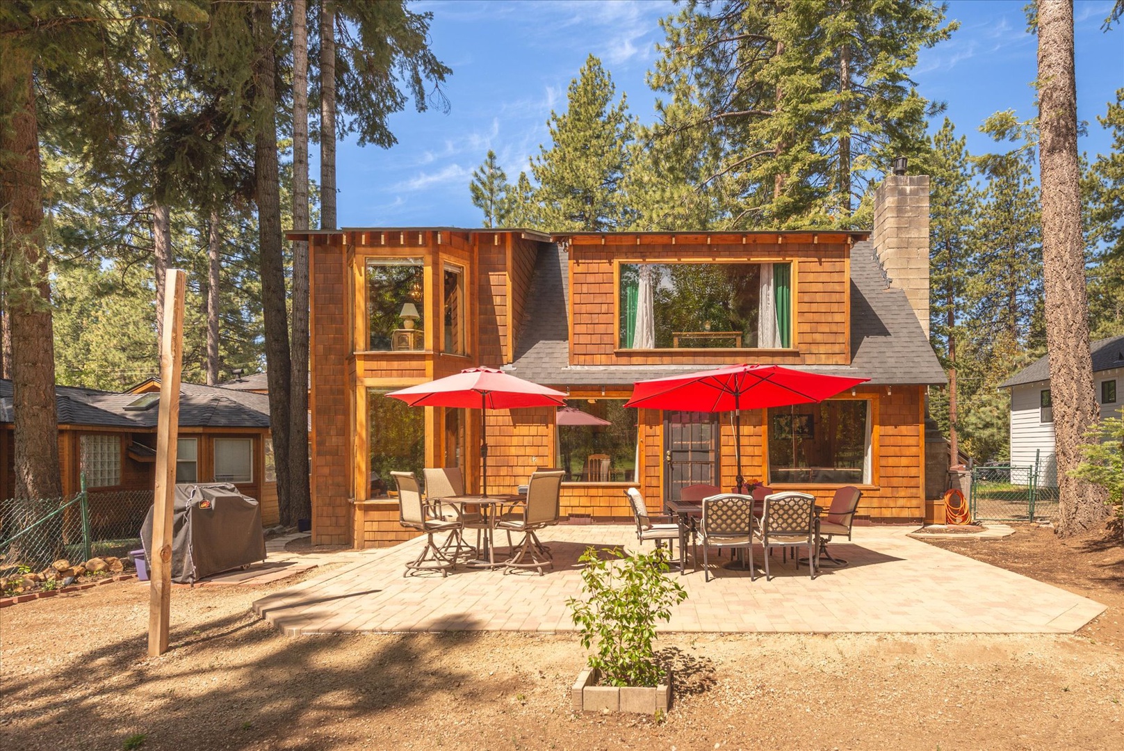 A charming cedar cabin nestled among towering pines, featuring a welcoming back patio with outdoor dining and bright red umbrellas for perfect forest getaways.