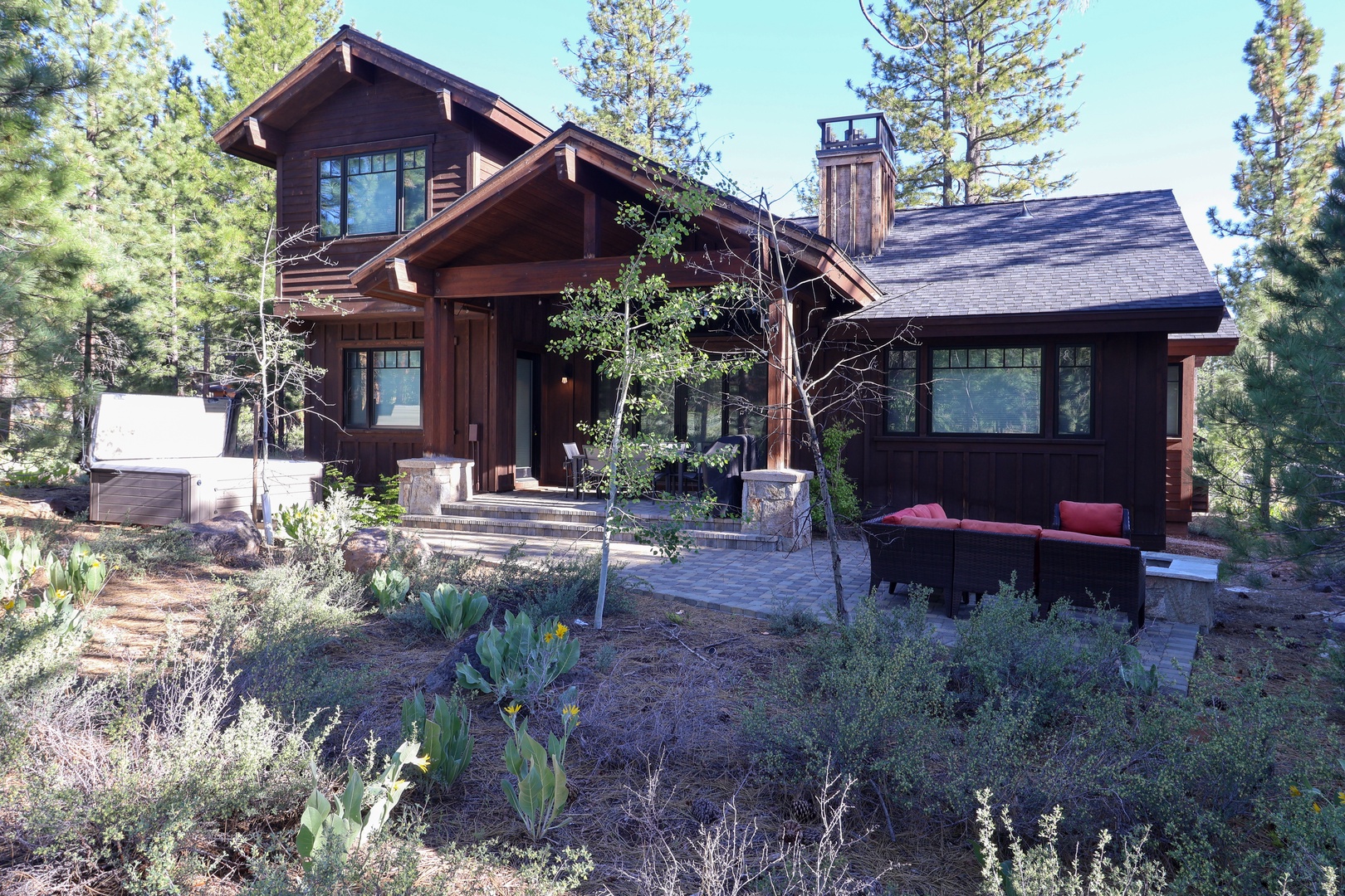 This stunning mountain lodge features a spacious patio with inviting seating, surrounded by towering pines and native landscaping, offering a serene arrival experience to your alpine getaway.