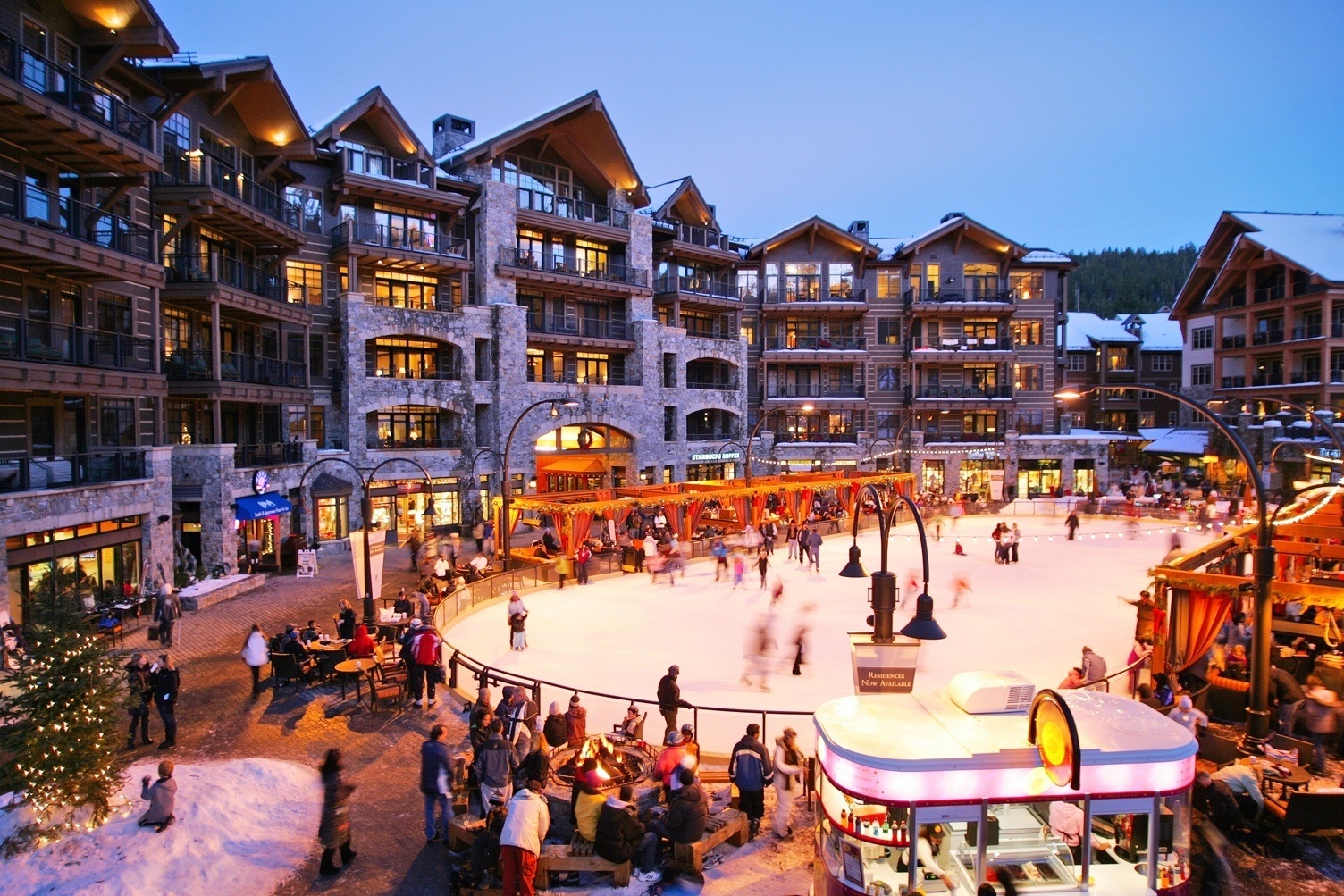 A vibrant mountain resort village plaza with ice skating, surrounded by alpine architecture and glowing with evening warmth.