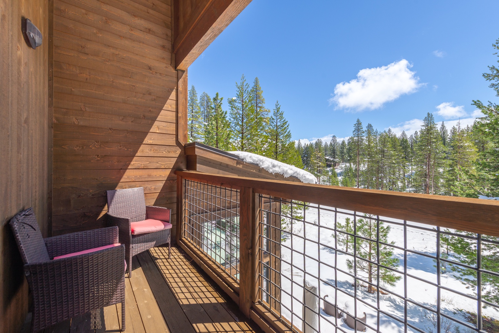 Wooden cabin balcony with comfortable seating overlooking snow-dusted evergreen forest beneath clear blue skies.