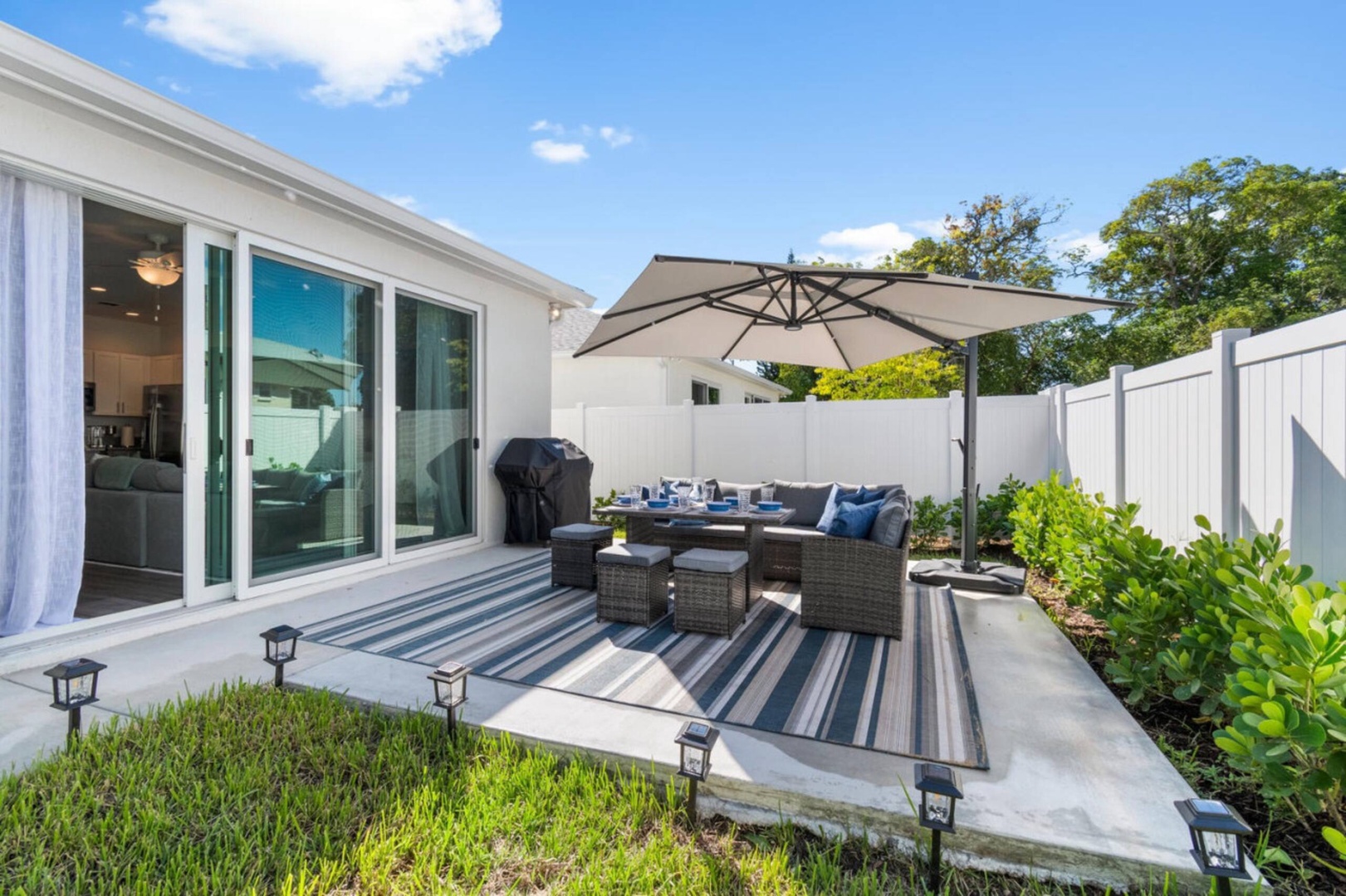 A bright and inviting outdoor patio awaits you, complete with comfortable seating, a dining area, and a BBQ grill, all framed by lush greenery and sunny skies.