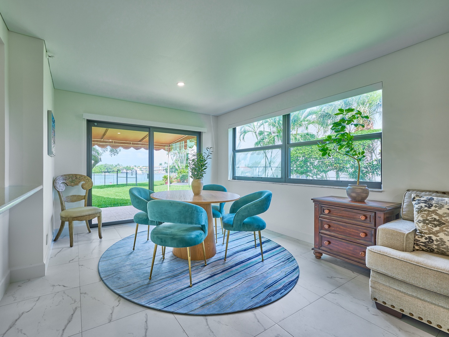 Step into your bright dining nook where plush teal chairs await your morning coffee. Large windows frame lush greenery and the patio beyond, inviting you to enjoy seamless indoor-outdoor living.