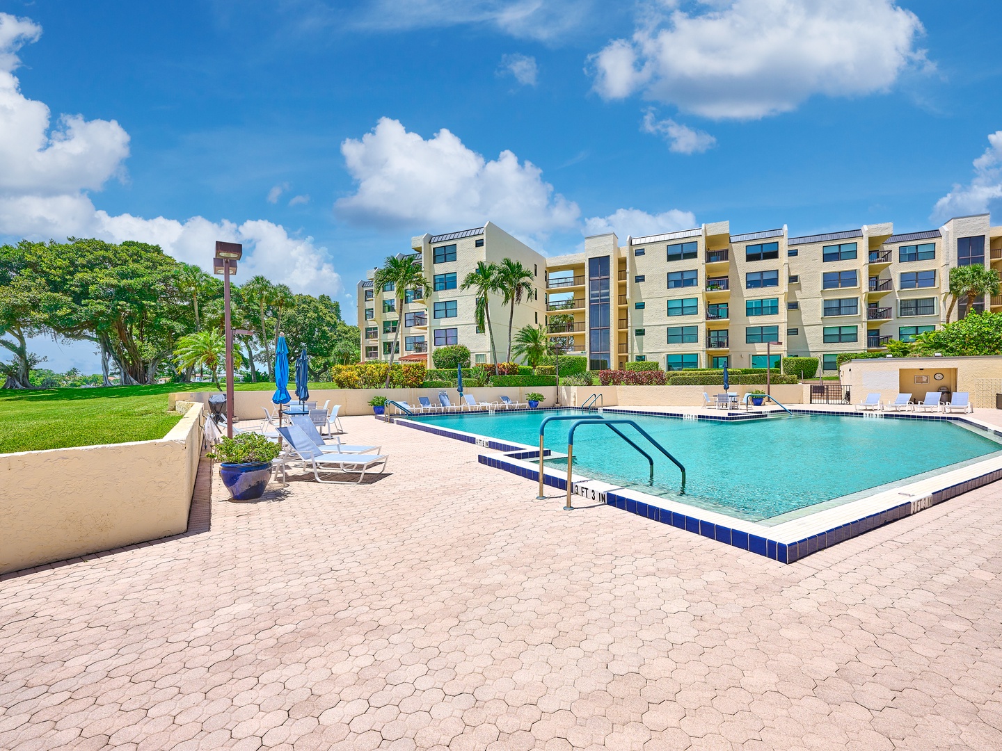 Modern waterfront property featuring a spacious pool surrounded by tropical landscaping and palm trees, set against vibrant blue skies.