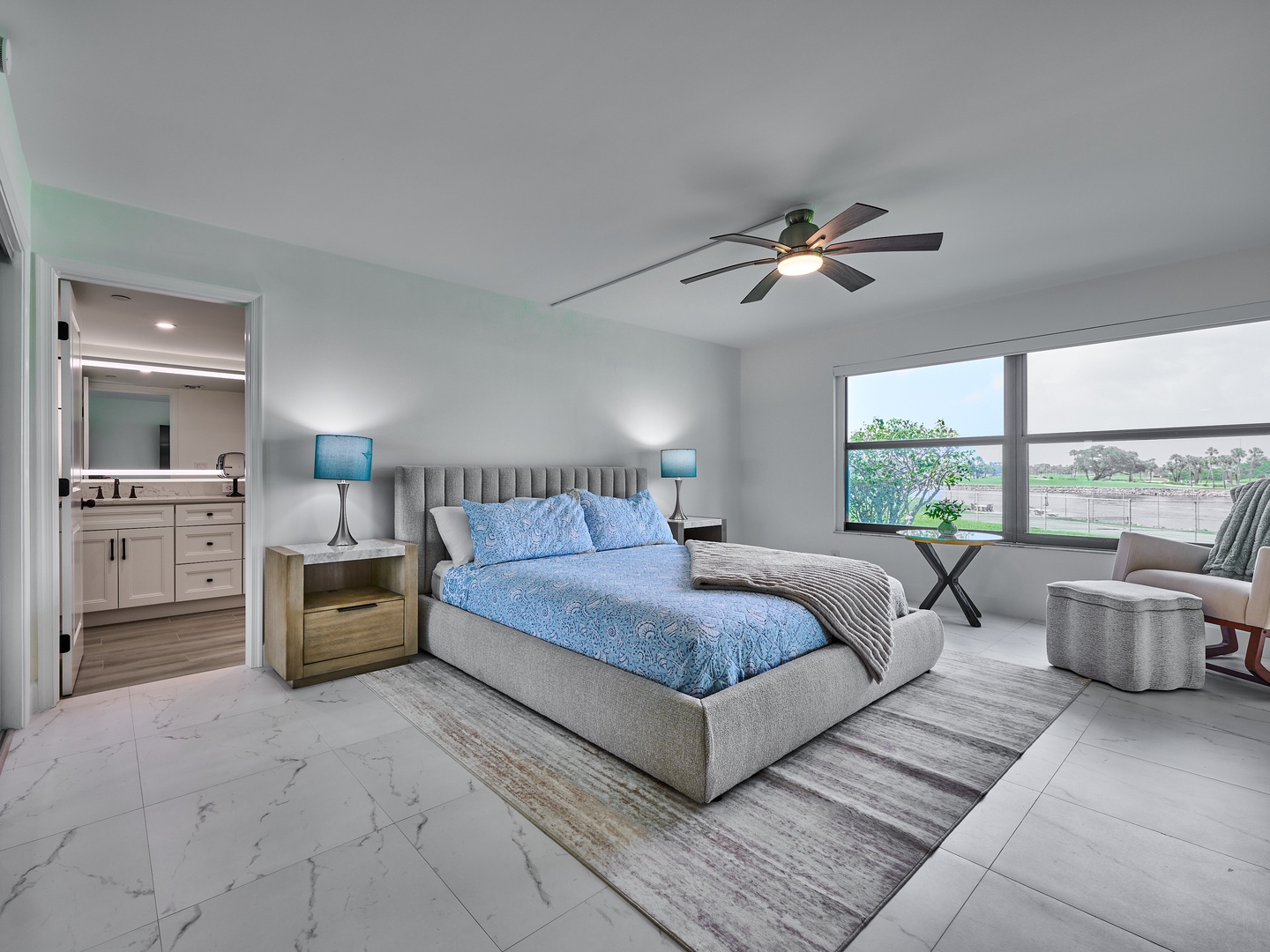 Drift off to sleep in your spacious master suite with soothing blue accents, a cozy upholstered bed, and gentle natural light streaming through oversized windows that frame views of greenery beyond.