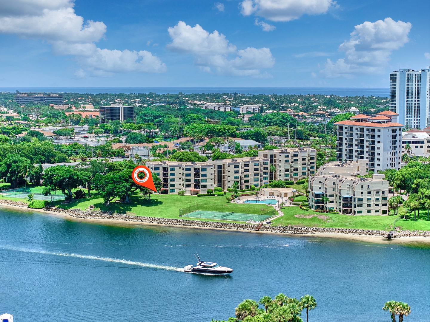 Aerial view of a coastal property complex surrounded by tropical greenery, with a boat cruising the nearby waterway.