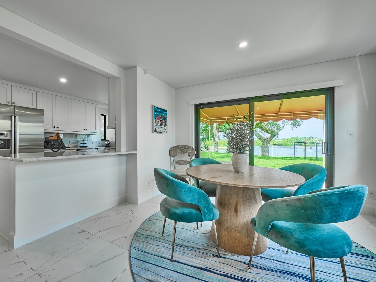 Bright dining space with vibrant turquoise chairs overlooking the open kitchen. Glass doors frame lush greenery and a covered patio—perfect for relaxed meals and easy entertaining.