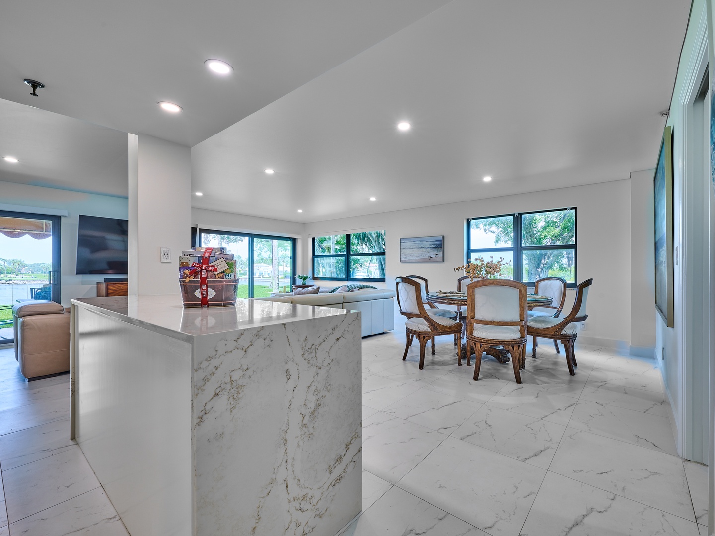 Natural light floods this open living space with a modern kitchen anchored by a marble island, flowing seamlessly into the dining area. Large windows frame lush greenery outside.