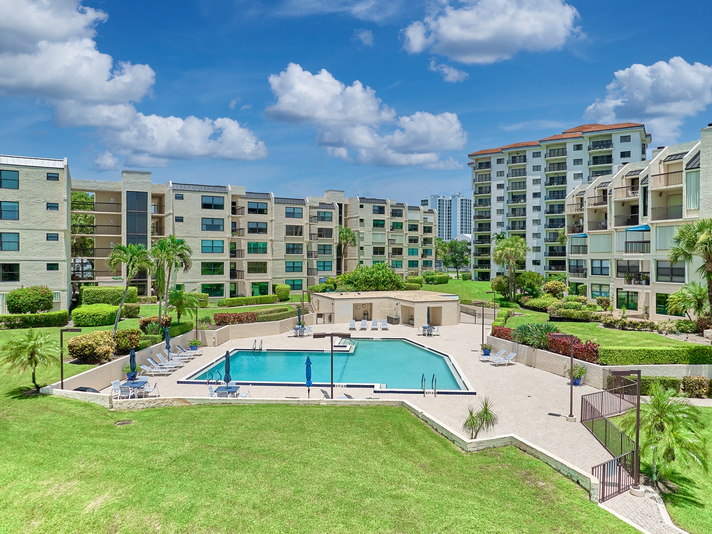 A tropical retreat featuring a sparkling pool surrounded by manicured lawns, palm trees, and lush landscaping. The complex is nestled within a peaceful residential area with modern buildings nearby.