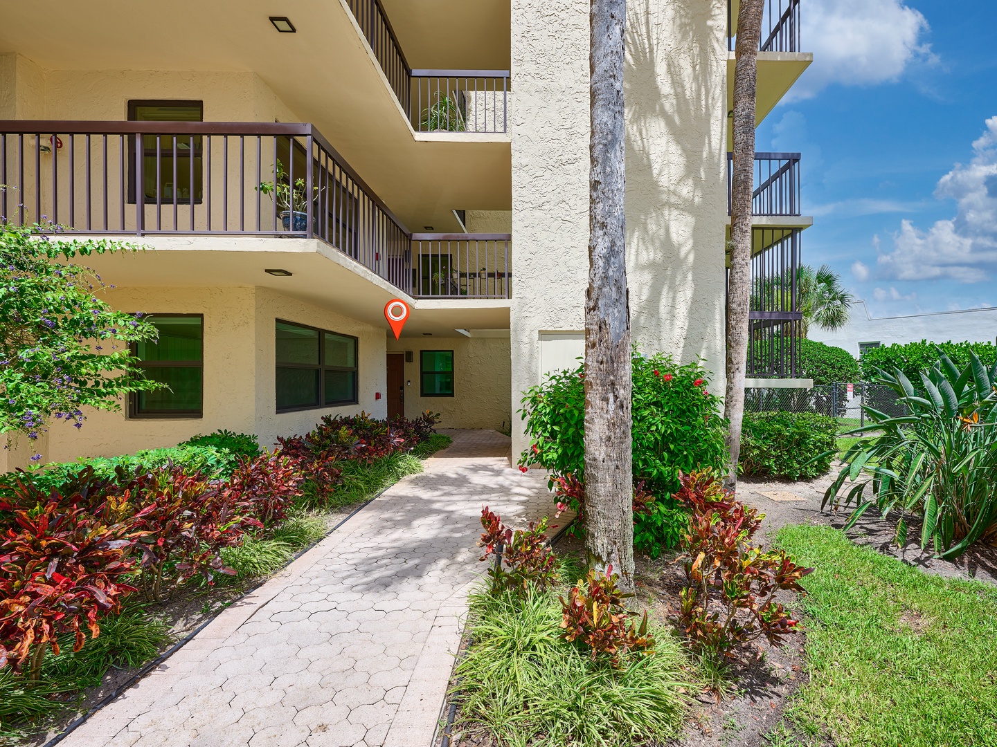 A multi-level building surrounded by lush tropical landscaping with colorful foliage, palm trees, and a paved walkway under sunny skies.