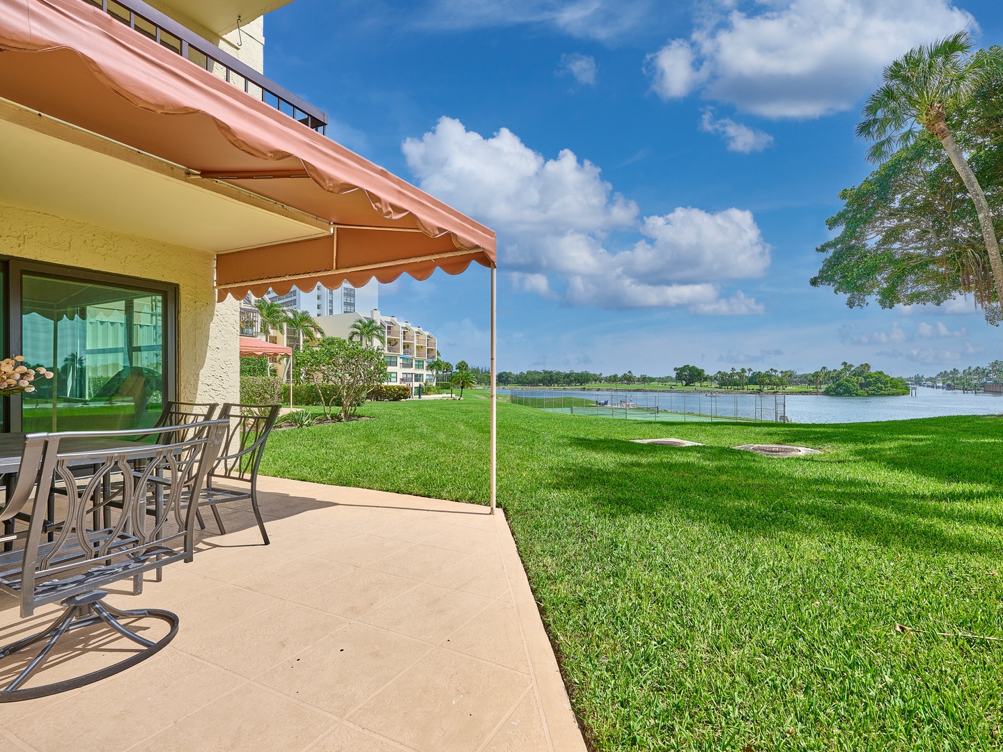 The property offers a peaceful lakefront location with lush green lawns and a private patio shaded by an awning, perfect for outdoor dining with tranquil water views.