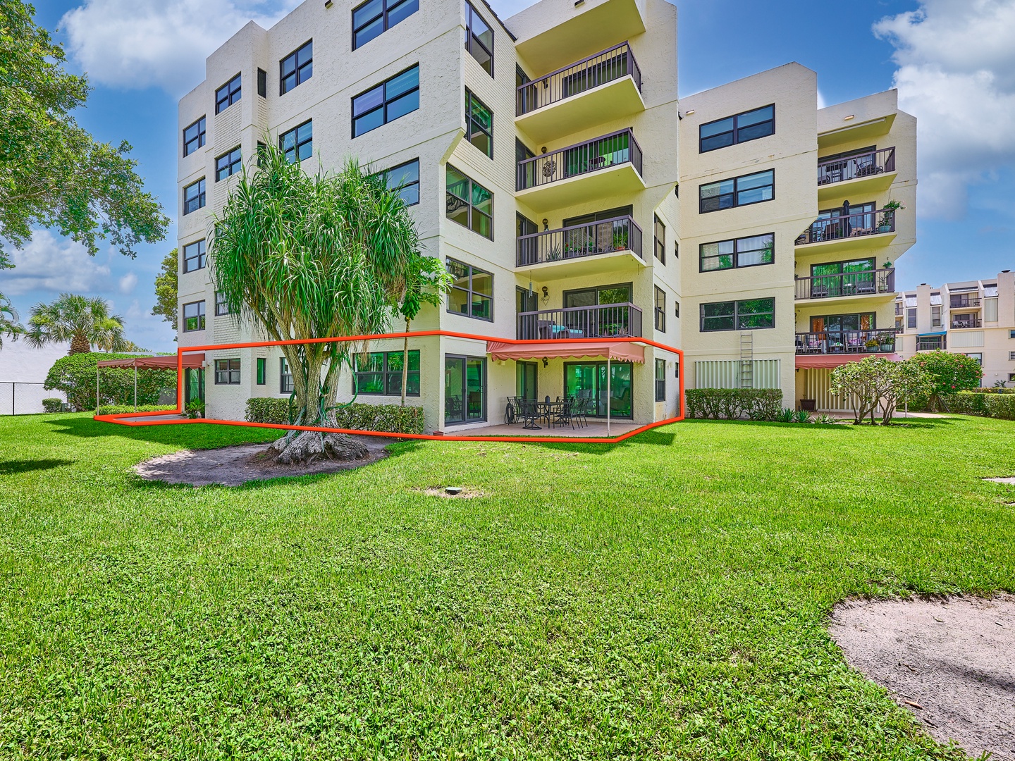 Modern multi-story building features a spacious private patio and lush lawn area surrounded by tropical landscaping. Ground-floor access offers seamless indoor-outdoor living with vibrant balconies above.