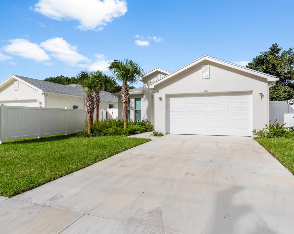 This modern Florida home features a welcoming facade with contemporary design, lush tropical landscaping, and a two-car garage, offering a warm arrival experience in a peaceful neighborhood.
