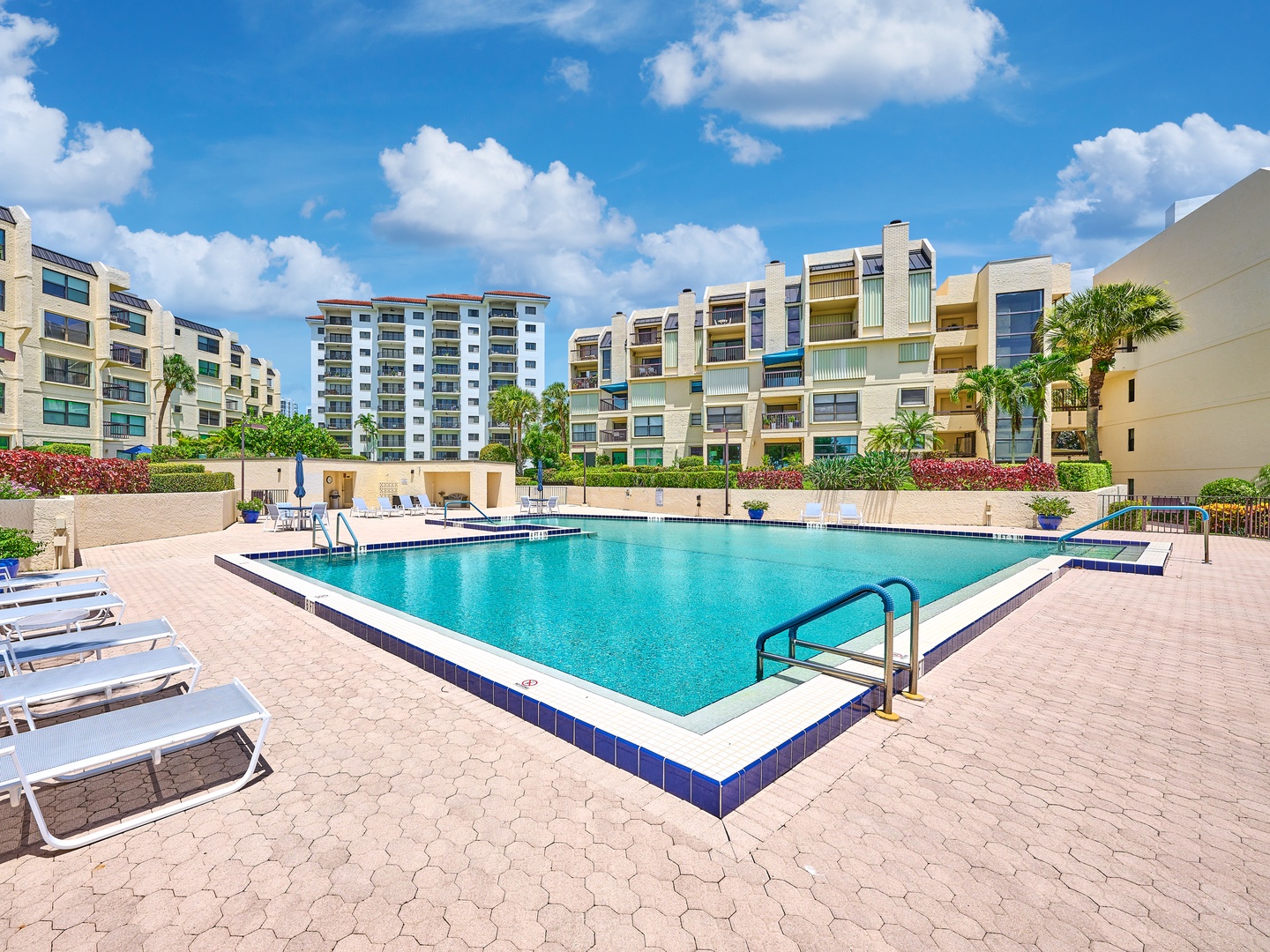 Expansive outdoor pool surrounded by comfortable lounge chairs and tropical landscaping with palm trees and vibrant flowers.