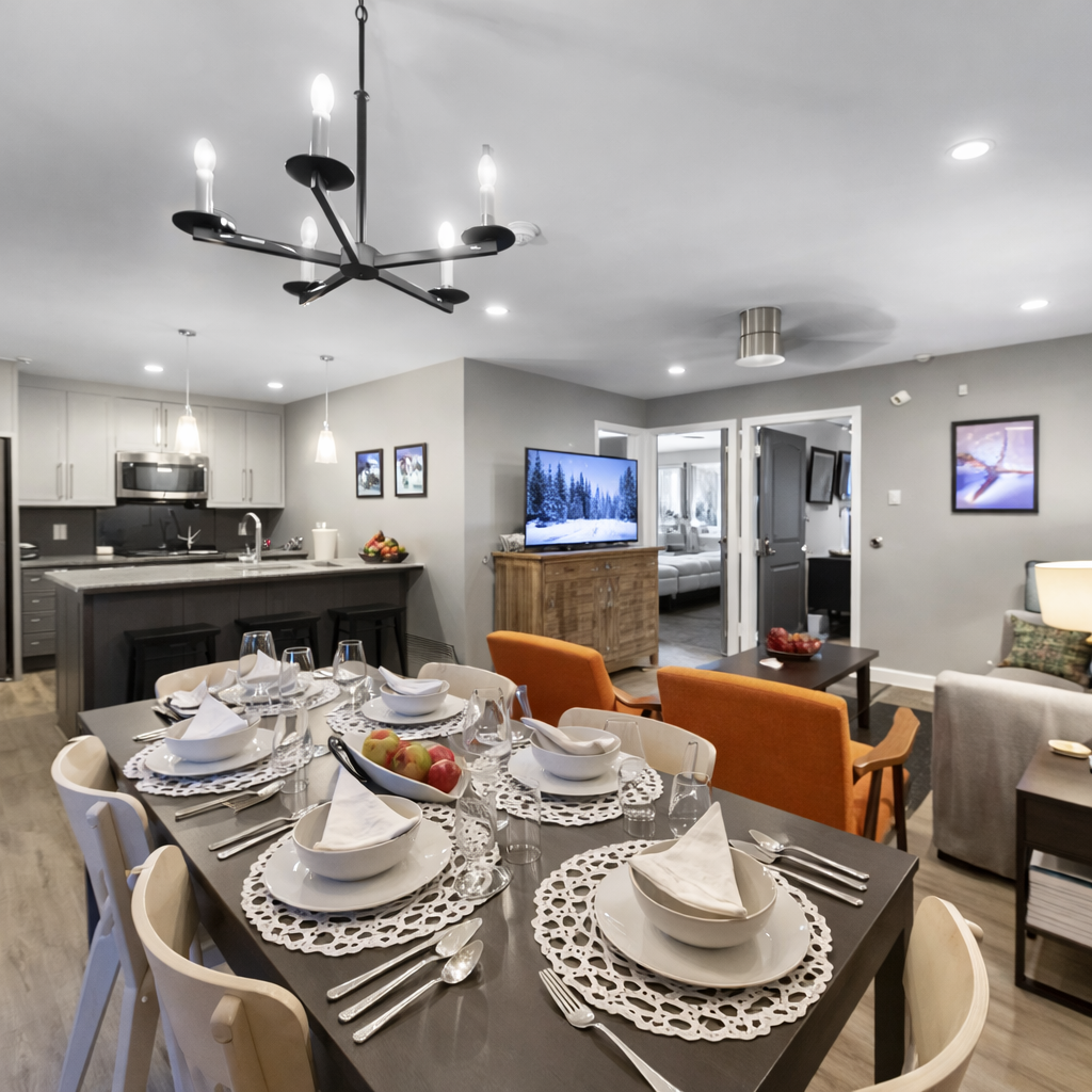 Bright, modern open-concept space with a fully set dining table, cozy seating, and a snowy-scene smart TV. Warm lighting and stylish décor create an inviting après-ski atmosphere.
