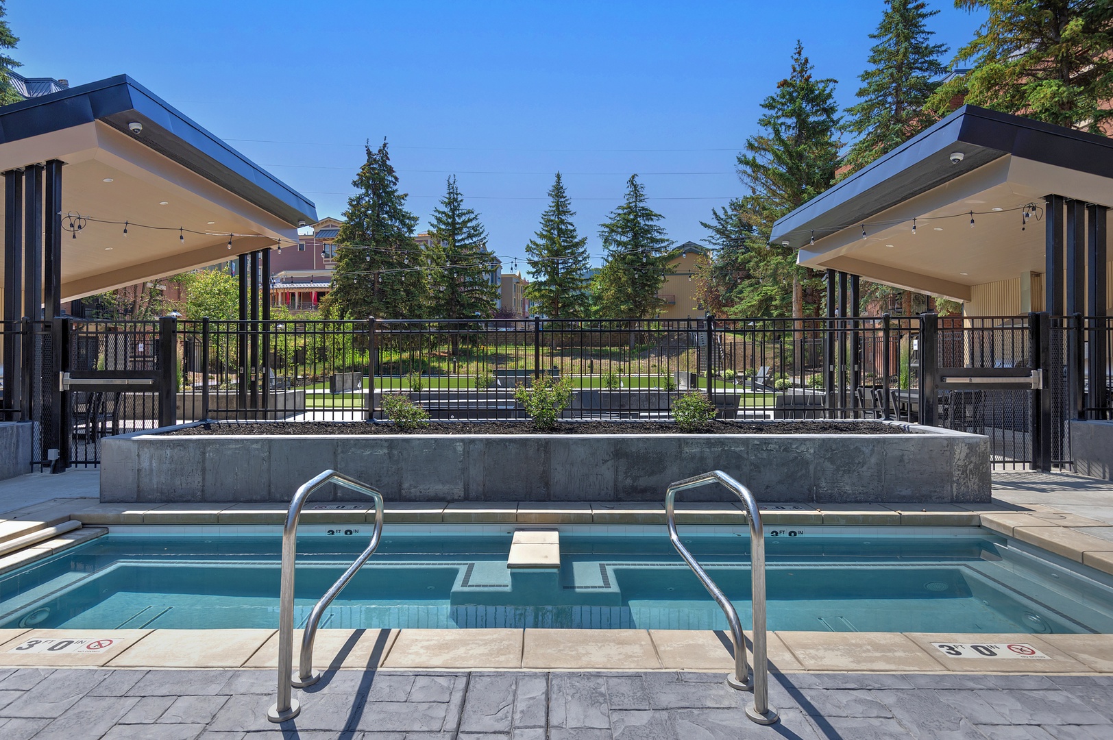Slip into clear turquoise waters and unwind at your private poolside cabana, where modern design meets relaxation. Two sleek pavilions offer shaded seating while evergreens frame peaceful views beyond the fence.