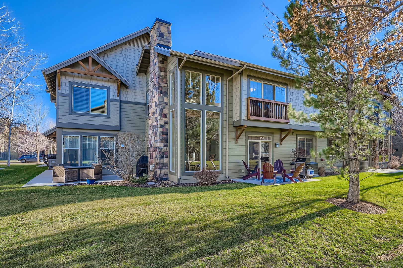 A charming two-story retreat with a spacious lawn where children play soccer, adults relax on the patio, and soaring windows frame mountain views under a blue sky.