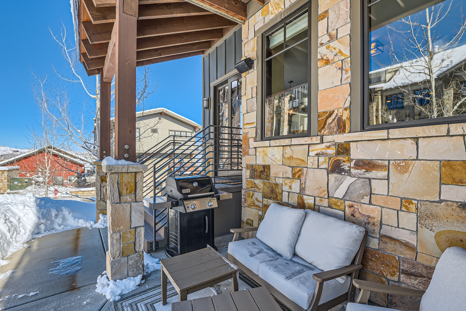 Winter welcomes you to a cozy outdoor retreat, where a covered patio offers comfortable seating, a BBQ ready for grilling, and a stunning mountain backdrop with snow-dusted trees and crisp blue skies.