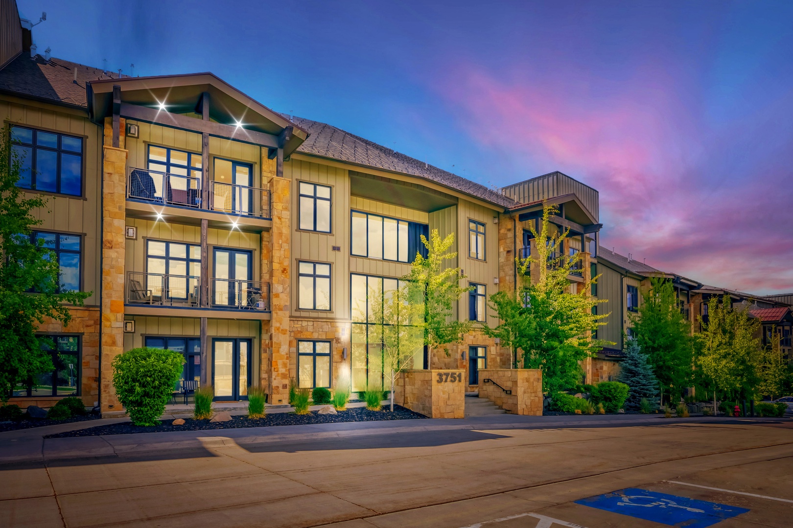 The property's exterior showcases modern mountain architecture with warm stone accents and elegant balconies, nestled among mature trees and landscaping with a stunning twilight sky overhead.