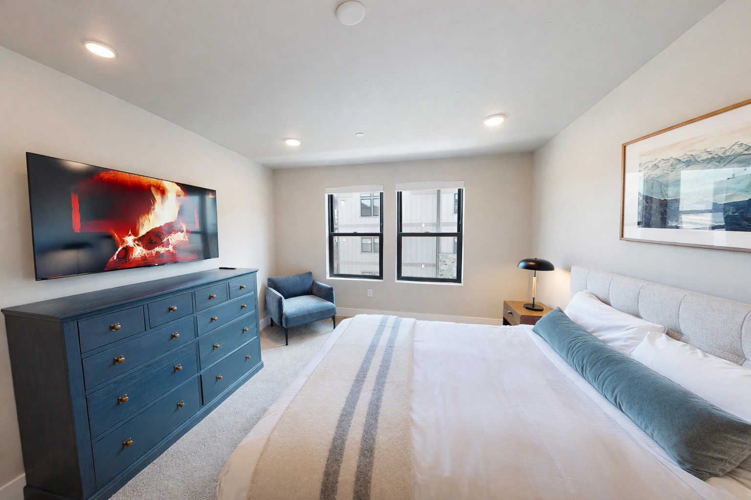 Stay in this serene, modern bedroom featuring a plush king-sized bed with a stylish channeled headboard. Large windows flood the space with light, while a cozy armchair offers the perfect spot to unwind.