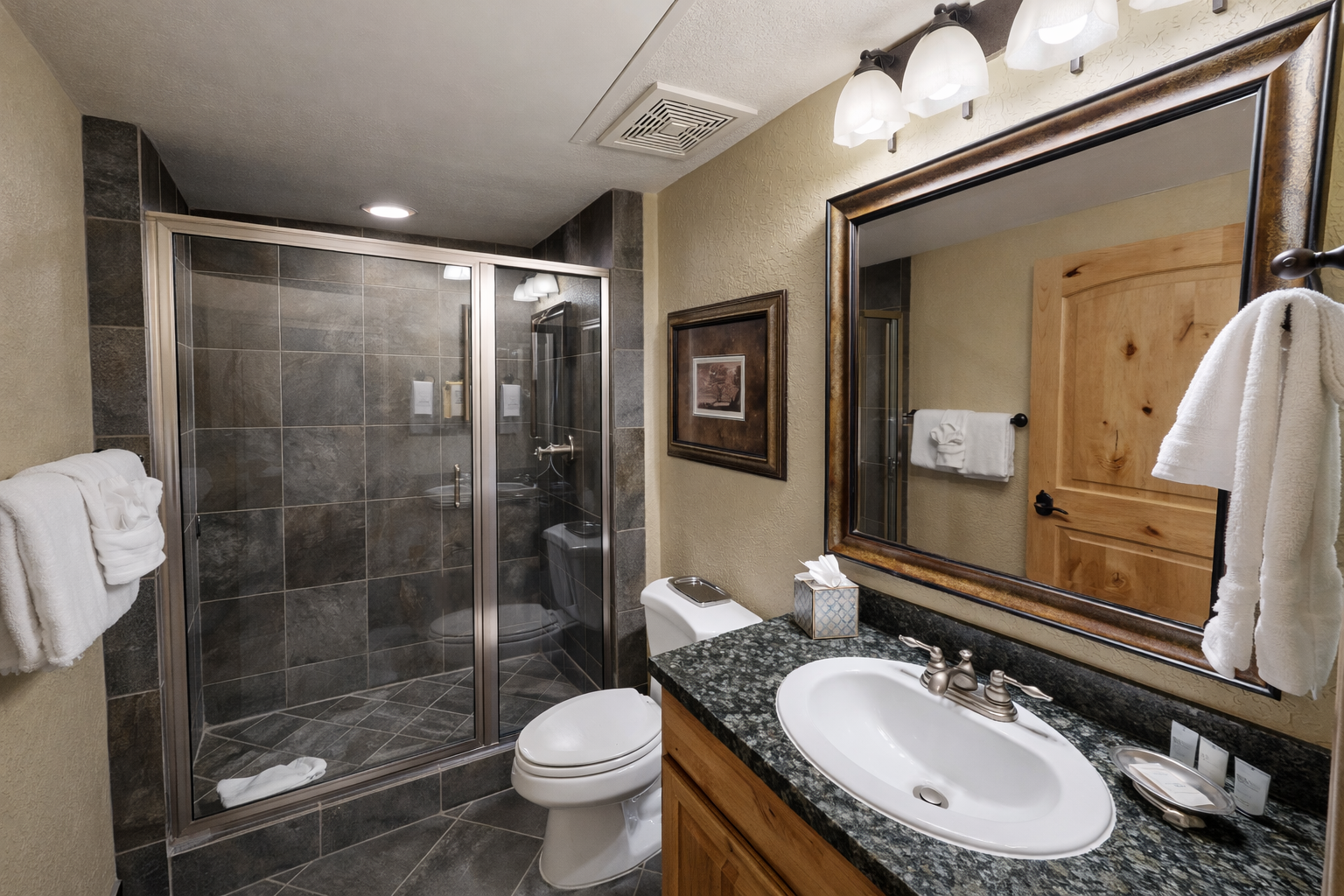 This modern bathroom features a sleek glass-enclosed shower with dark tile and a stylish granite vanity. Bright lighting and a large framed mirror create a clean, inviting space.