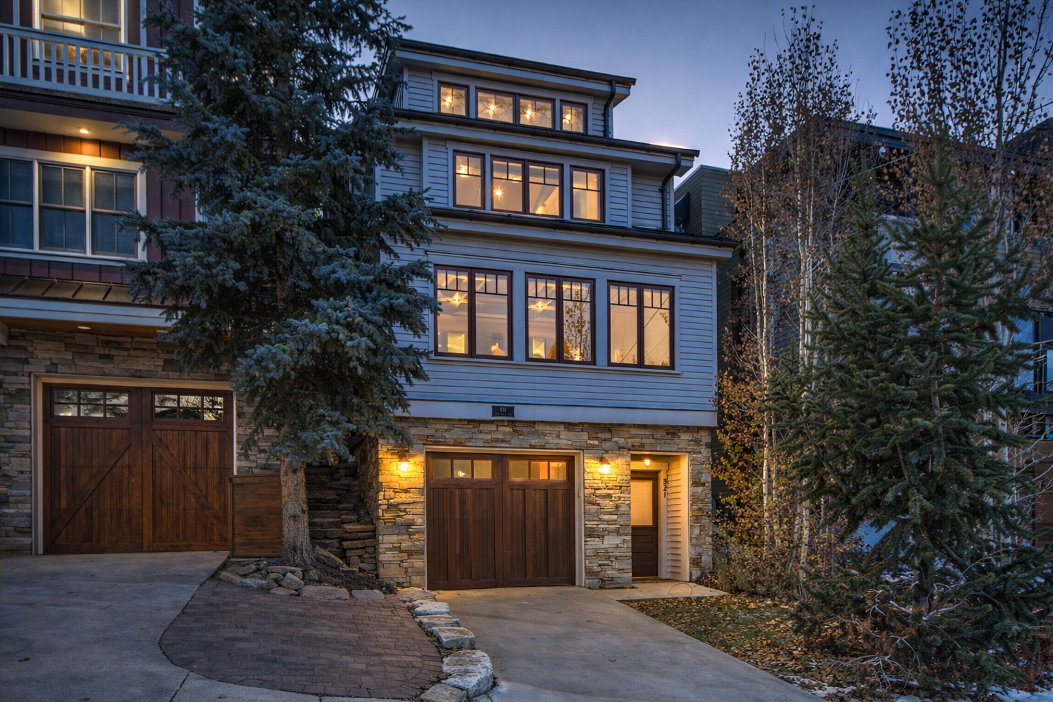 Modern mountain home with stone and siding exterior, illuminated by warm evening lighting among mature trees.