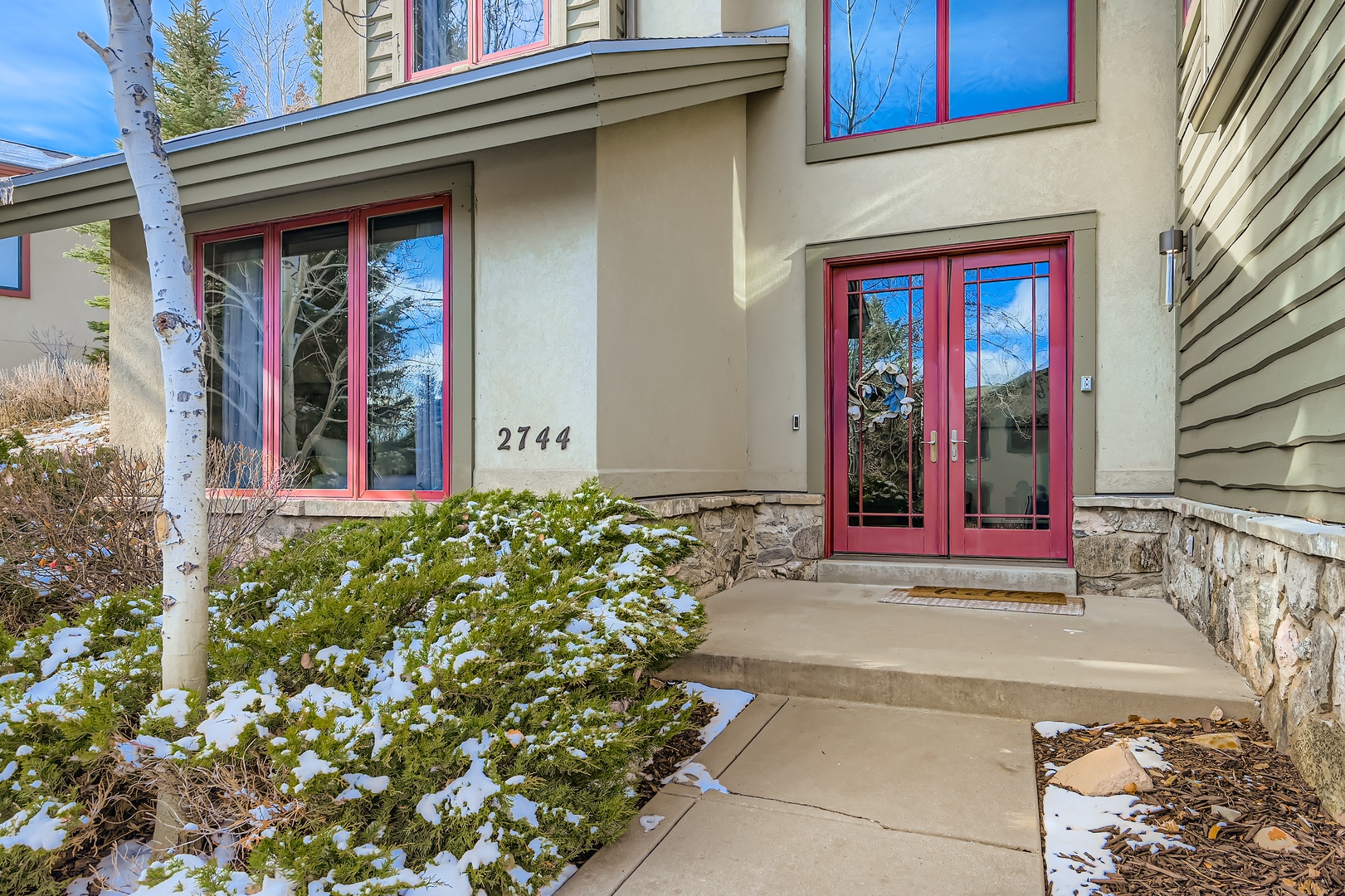 The welcoming entrance features a red-framed double door and number 2744, surrounded by natural stone accents and landscaped greenery dusted with winter snow—a quiet arrival moment in a beautiful mountain setting.