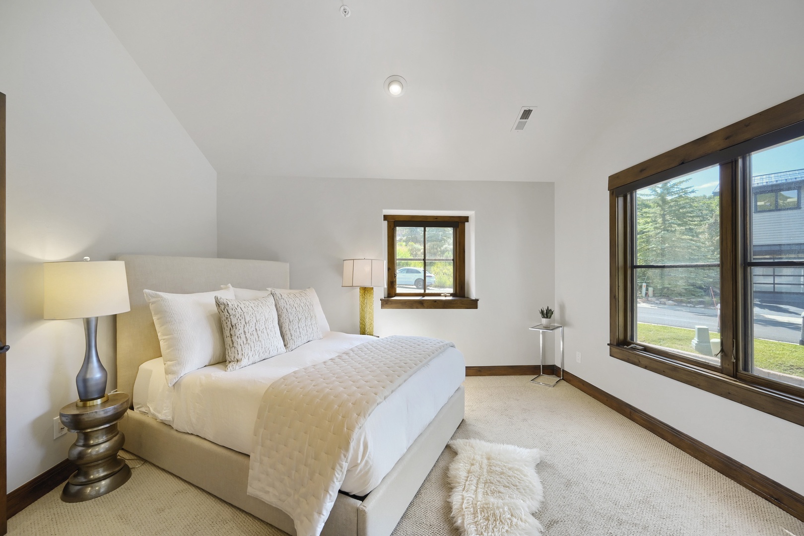 Your bright, restful bedroom features a cozy king bed dressed in soft linens, warm wood trim, and large windows that fill the space with natural light.