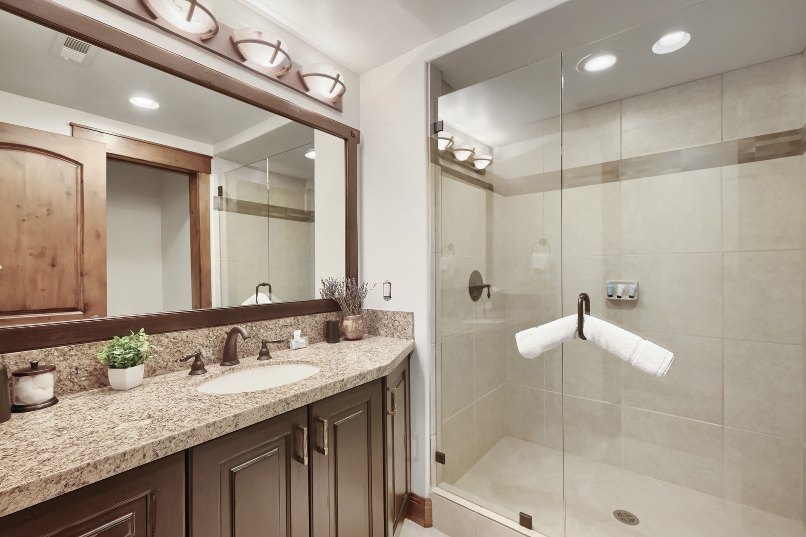Step into your private spa-like bathroom featuring warm wood tones, granite countertops, and a spacious glass-enclosed shower. Fresh plants and thoughtful details create a welcoming retreat for your stay.