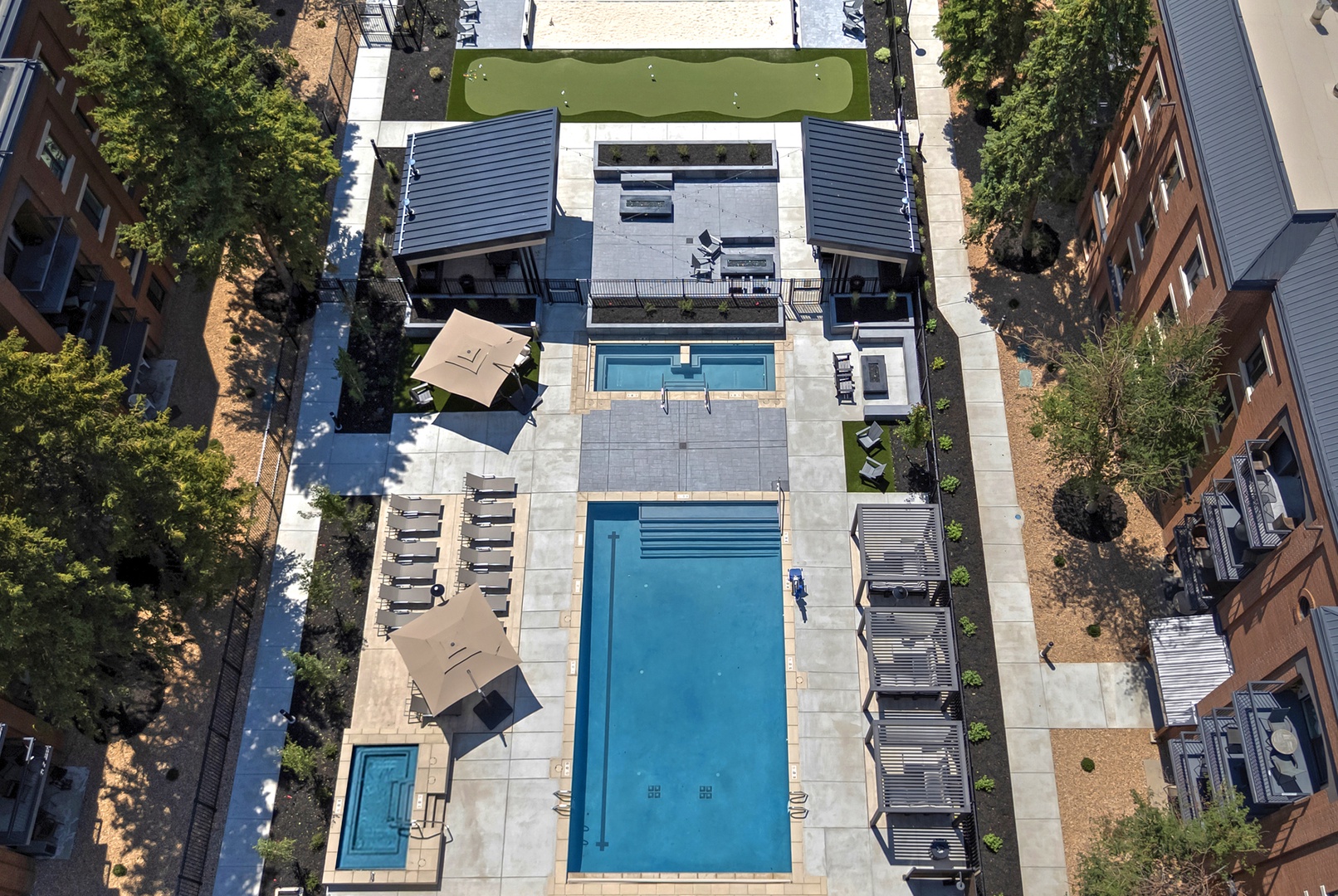 Aerial view showcasing modern outdoor amenities including dual swimming pools, sunbathing deck with umbrellas, spa, artificial turf lawn, and lounge seating—all within a secure, landscaped courtyard surrounded by residential buildings.