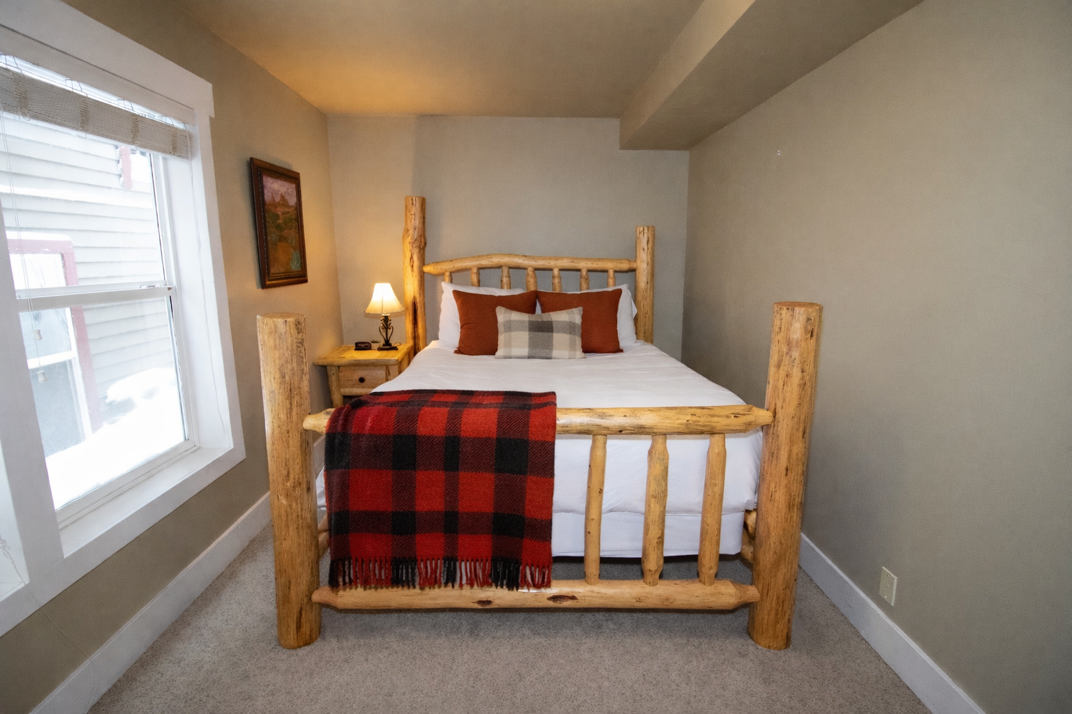 This charming secondary bedroom features a handcrafted log bed frame and cozy buffalo plaid accents. Large windows fill the space with natural light, creating a warm, inviting atmosphere for guests.