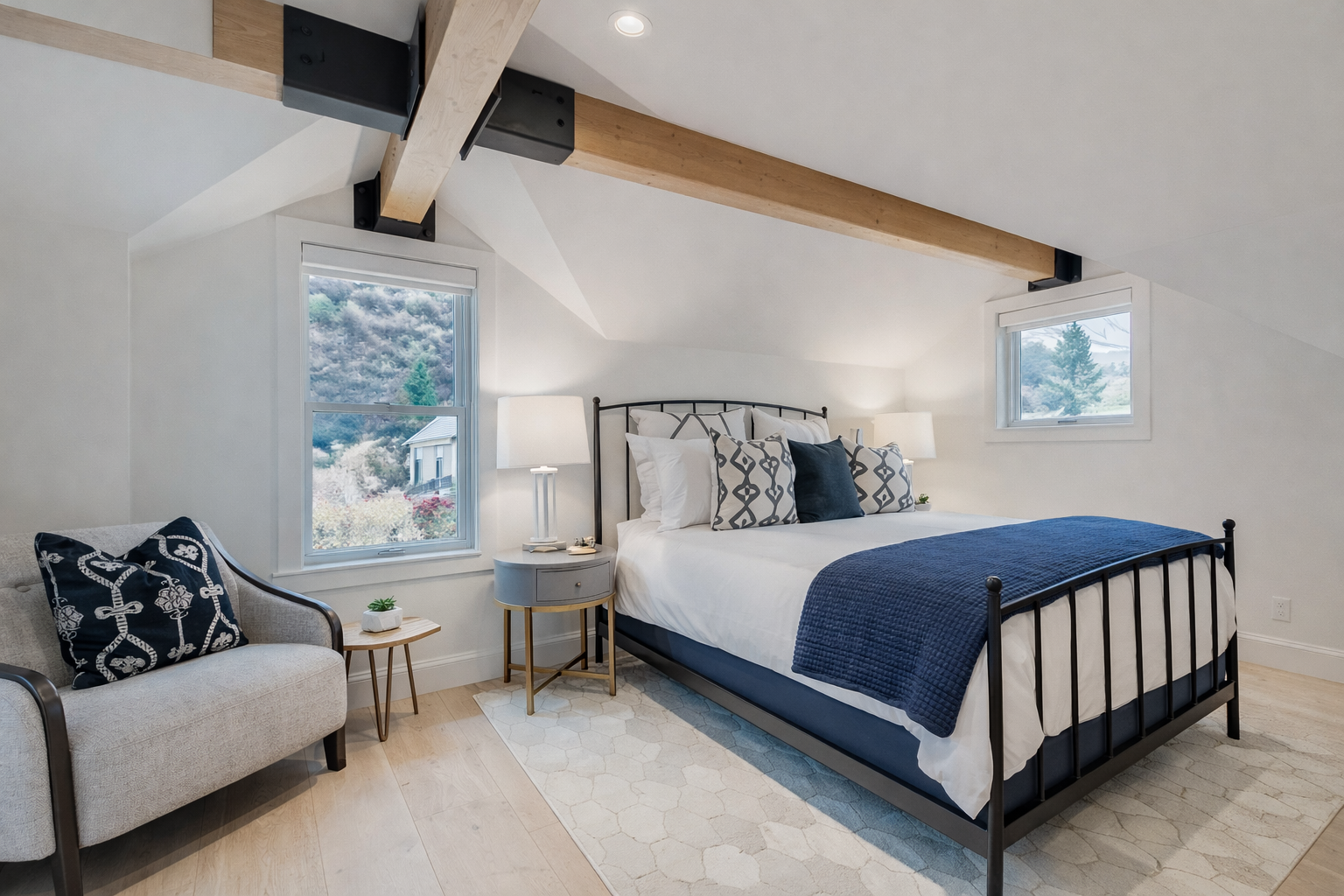 Unwind in your airy loft bedroom where natural light streams through dual windows, highlighting exposed wooden beams and cozy blue accents.