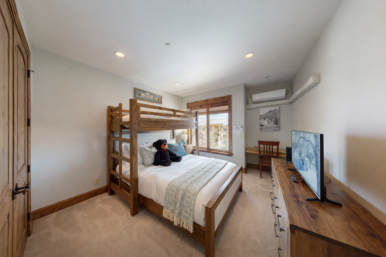 This versatile guest bedroom features a twin-over-queen bunk bed, perfect for families. Highlights include a dedicated workspace, large flat-screen TV, and ample storage, all in a bright, modern mountain setting.