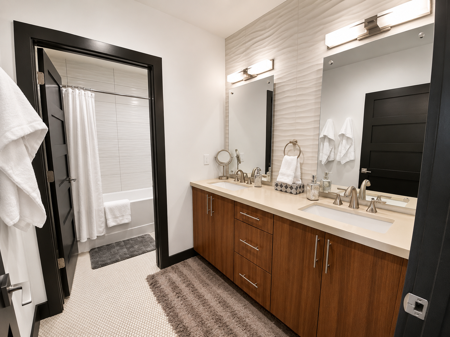 This modern ensuite bathroom features a spacious double vanity with wood-grain cabinetry and a private water closet with a soaking tub. Sleek mirrors and warm lighting create a bright, inviting space.