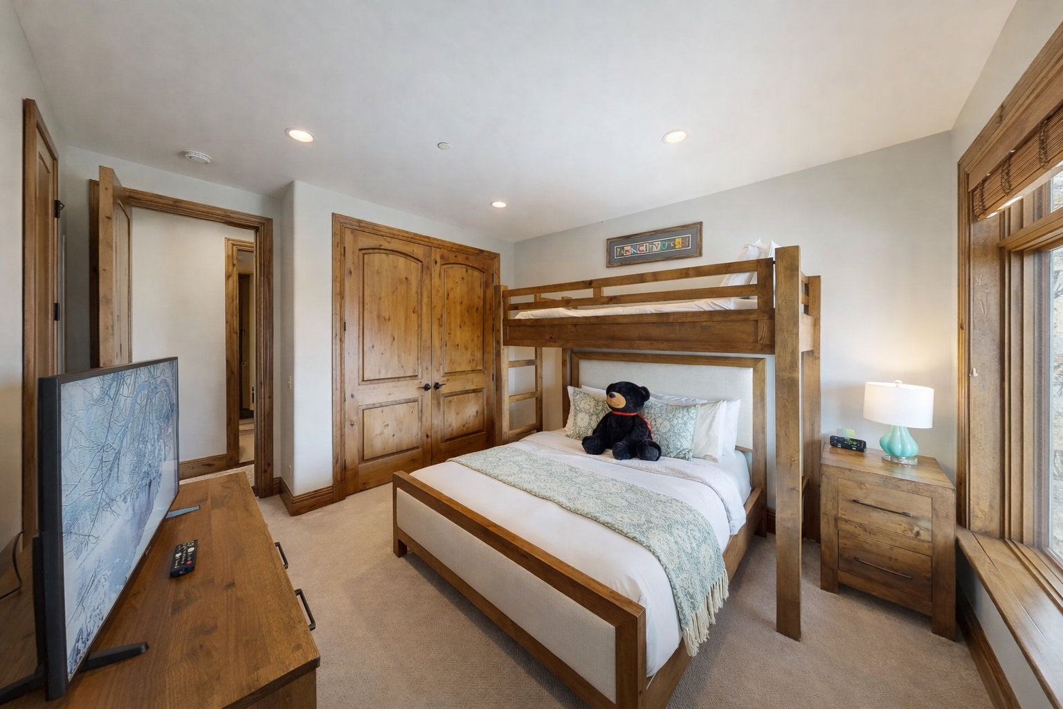 This versatile guest bedroom features a twin-over-queen bunk bed, perfect for families. Highlights include a dedicated workspace, large flat-screen TV, and ample storage, all in a bright, modern mountain setting.