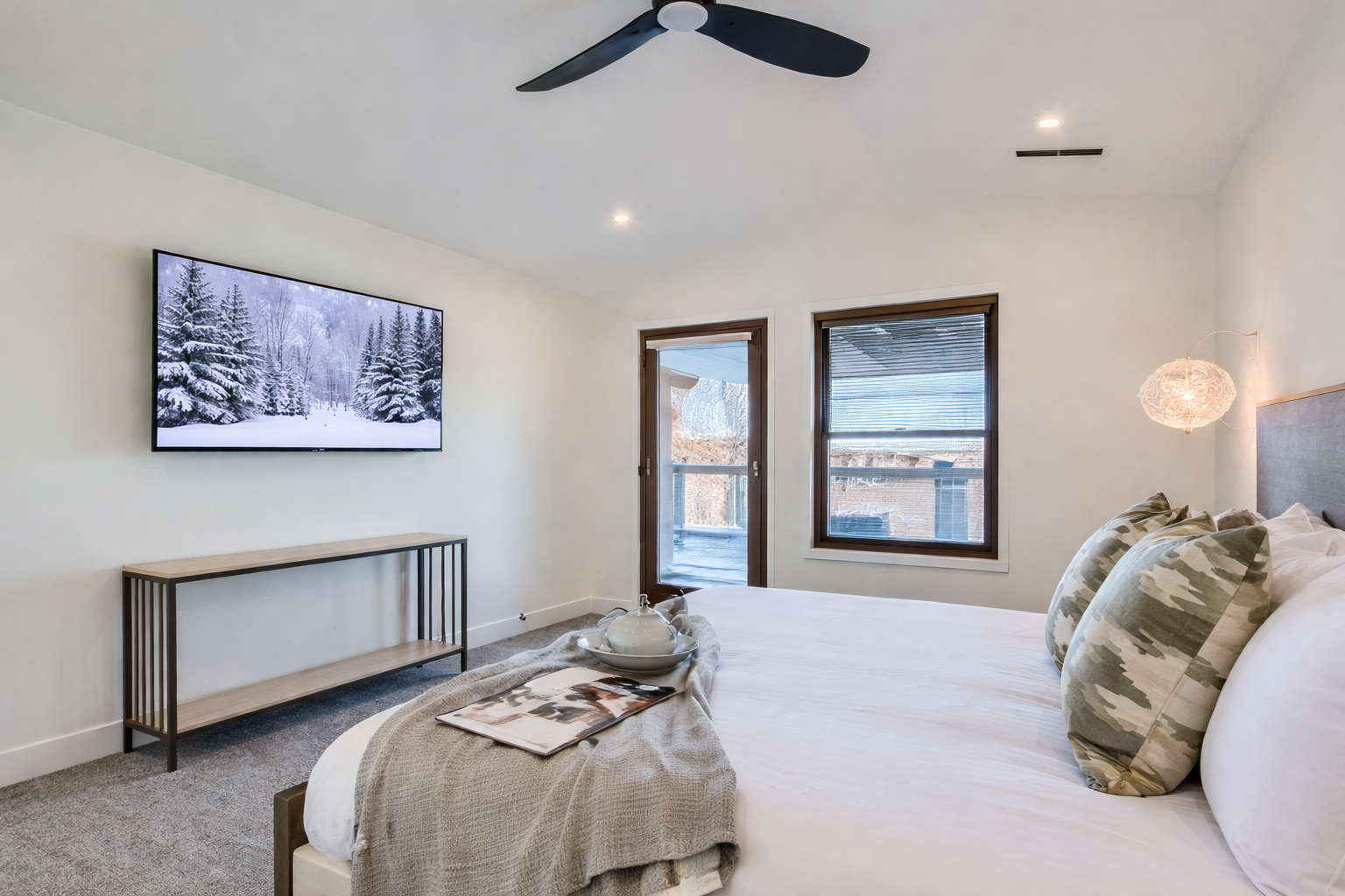 Bright, modern bedroom featuring a plush bed with crisp white linens, soft accent pillows, and a cozy throw. Large windows, balcony access, and a wall-mounted TV create a relaxing Park City retreat.