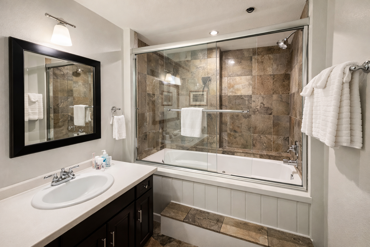 Modern & clean full bathroom featuring a sleek tub/shower combo with glass doors and stylish stone-look tile.