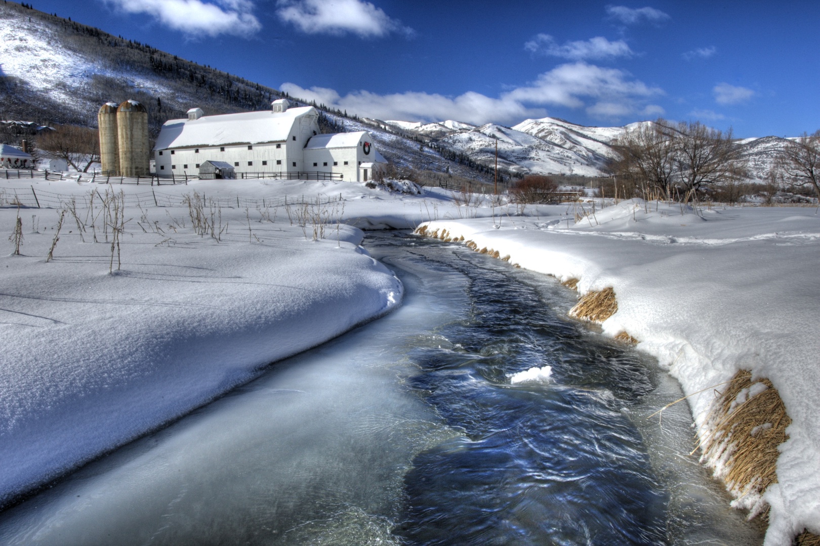 Snow-covered farm buildings nestled in pristine mountain valley with flowing creek and dramatic winter landscape.