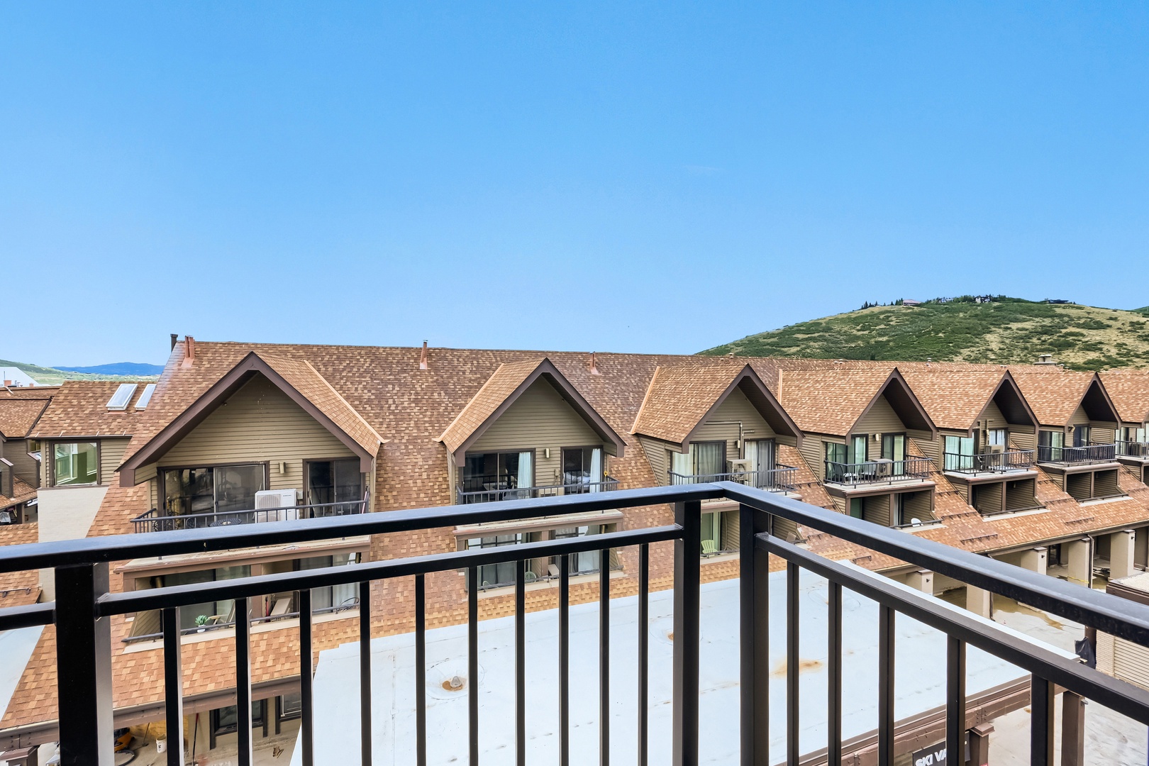 Charming mountain resort with chalet-style architecture set against rolling hillside terrain under clear blue skies. The property features distinctive peaked rooflines and warm earth-toned exteriors, surrounded by natural landscape.