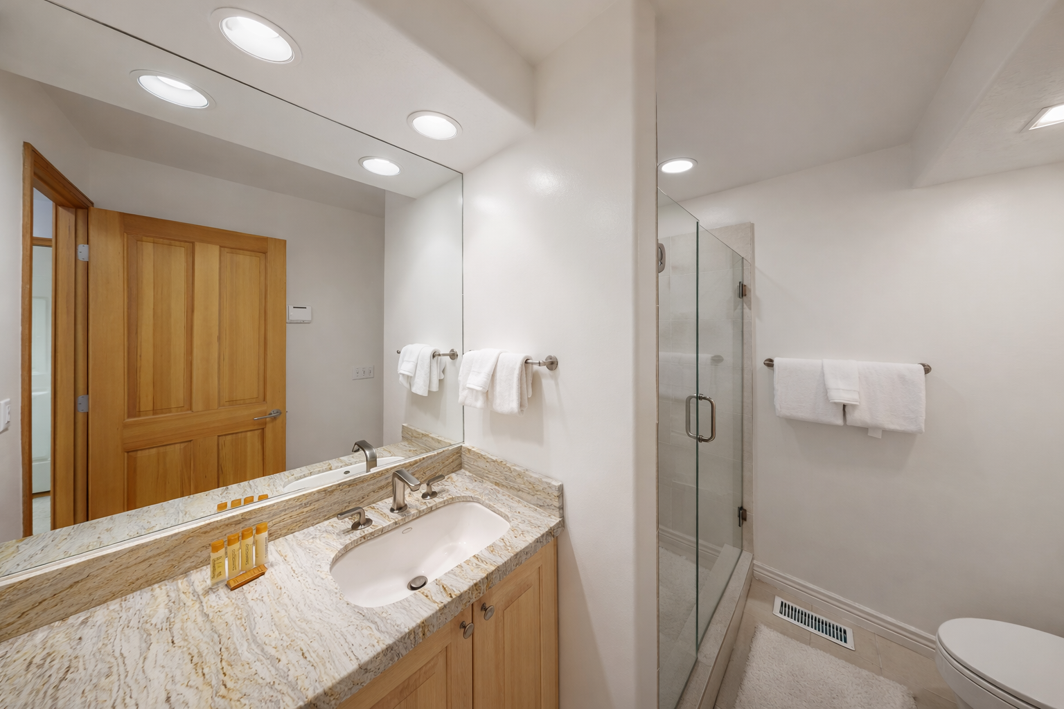 This modern bathroom features a spacious vanity with elegant granite countertops, a large illuminated mirror, and a sleek, glass-enclosed shower for your convenience.