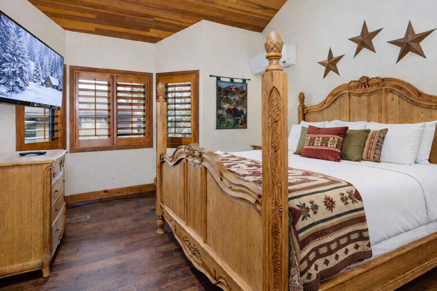 Relax in your spacious master bedroom featuring stunning wood beam ceilings, rustic furnishings, and modern amenities like AC and TV for the perfect mountain retreat.