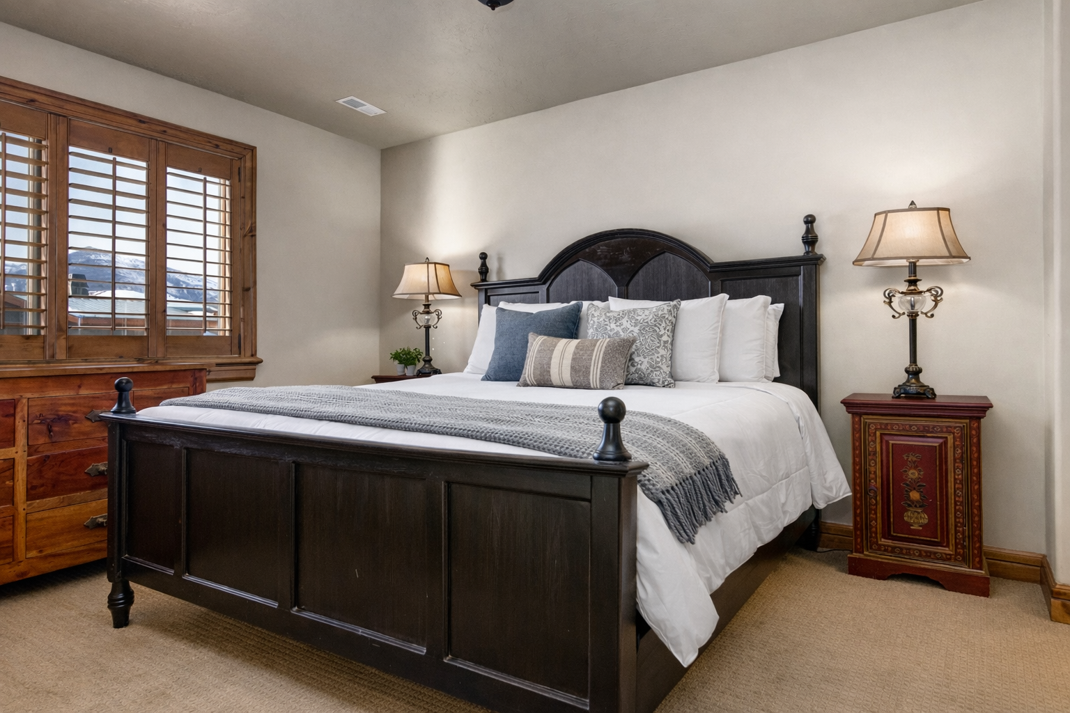 This spacious guest room features a grand four-poster bed, premium linens, and warm bedside lighting. Shuttered windows and plush carpeting create a quiet, comfortable retreat for a restful night.