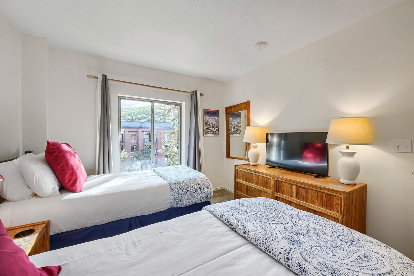 Your bright, airy bedroom features two cozy beds with crisp white linens and cheerful pops of color, plus warm lamps and a view of the surrounding neighborhood through your window.