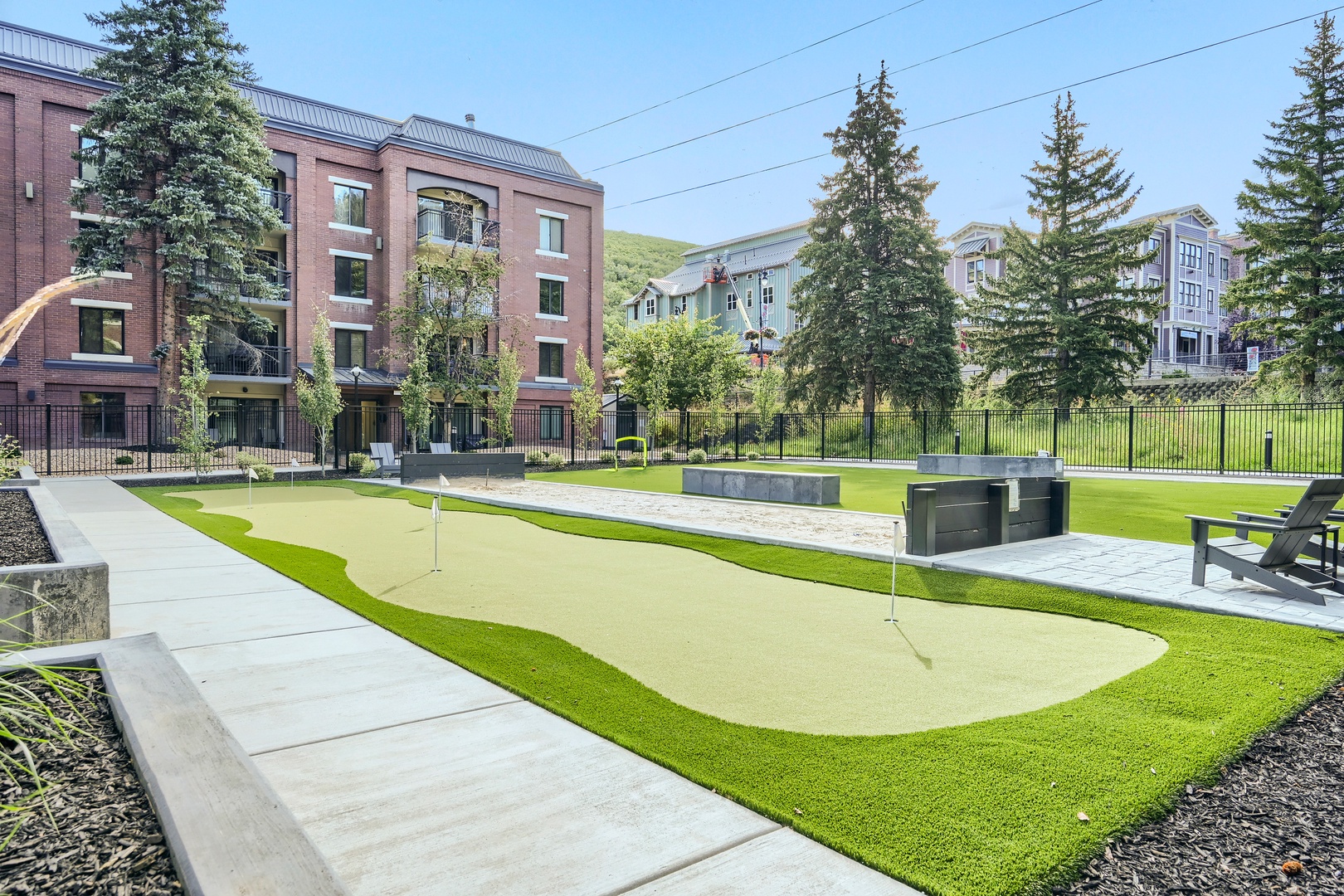 The property features a professionally designed putting green and outdoor recreational area, framed by mature evergreens and surrounded by modern residential buildings. Mountain backdrop adds natural beauty to the arrival experience.