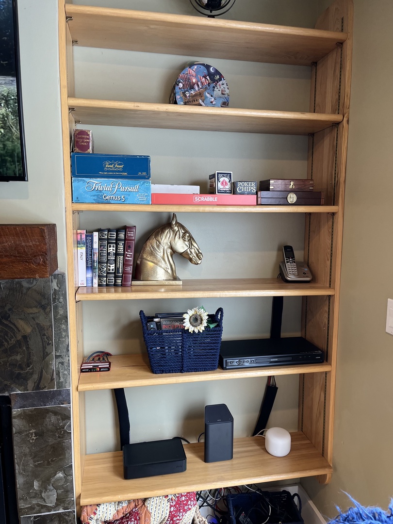 This light-toned wooden bookcase offers five tiers of organized storage. It currently displays a curated selection of board games, books, and unique decorative accents, including a striking brass horse head.